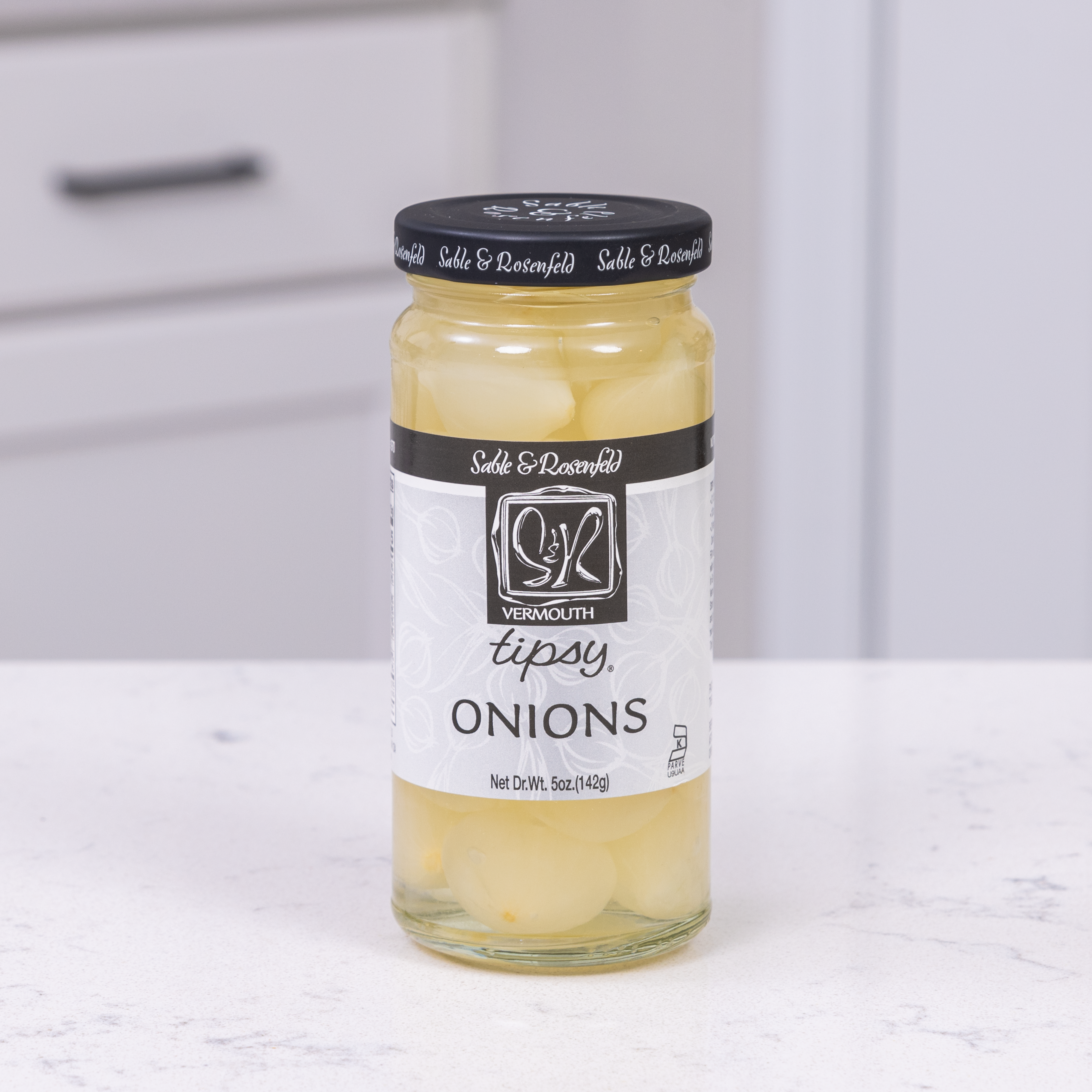 A jar of Vermouth Tipsy Onions by Sable & Rosenfeld, filled with silverskin pickled onions, placed on a white kitchen countertop with a modern cabinet in the background.