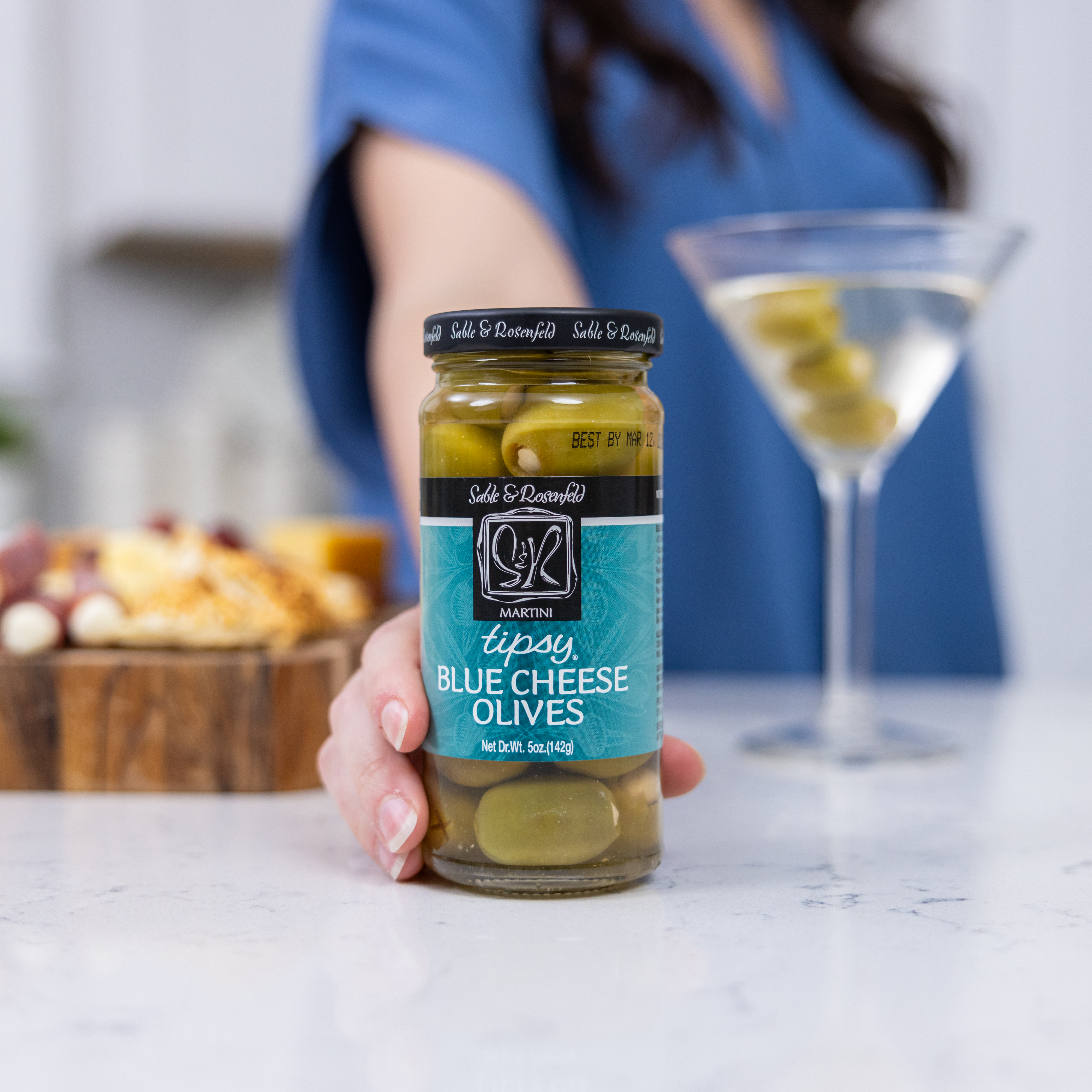 A jar of Sable & Rosenfeld Blue Cheese Tipsy Olives held in hand, with a martini garnished with olives and a charcuterie board in the background perfect for cocktail lovers.