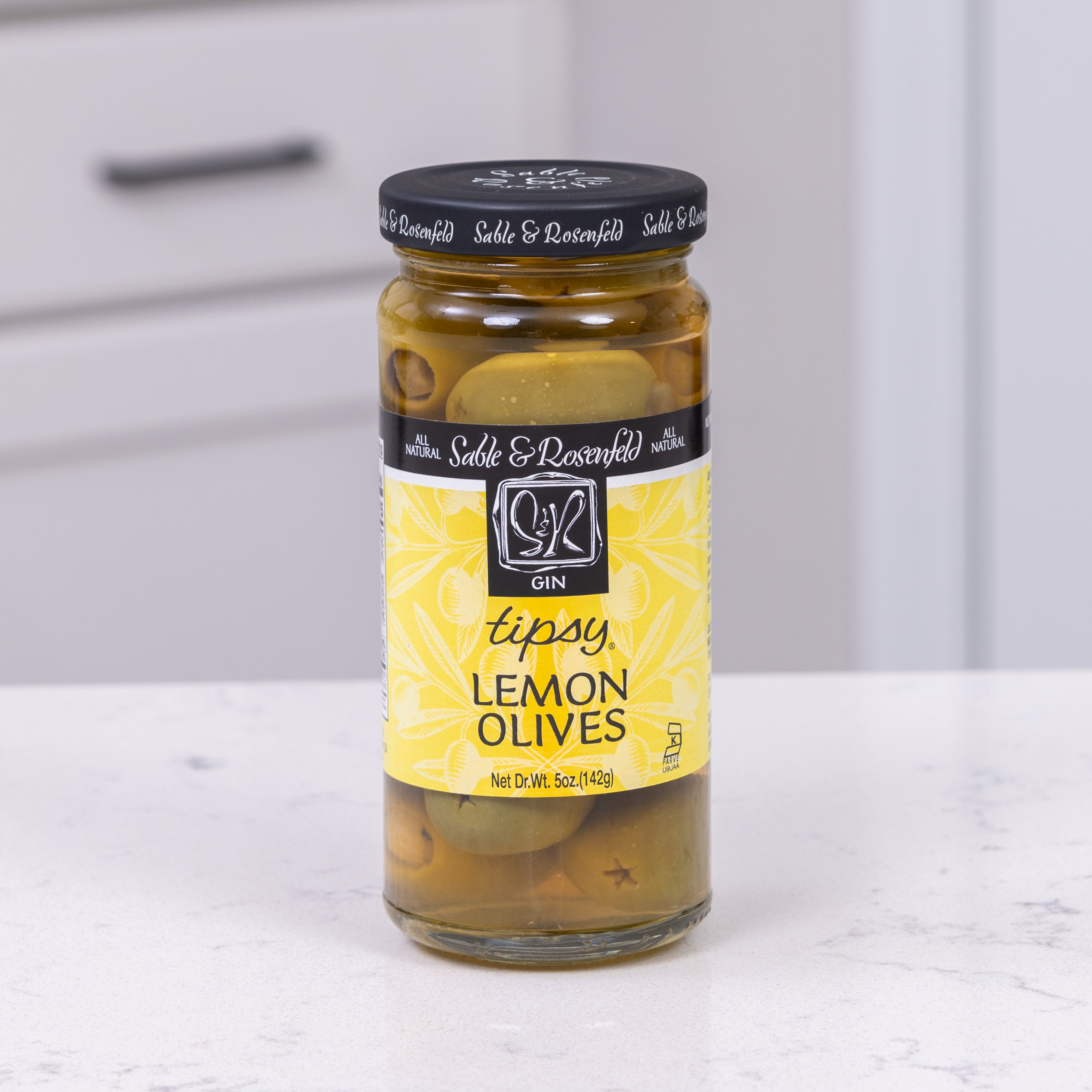 A jar of Gin Lemon Tipsy Olives on a kitchen counter, perfect for garnishing a Classic Martini.