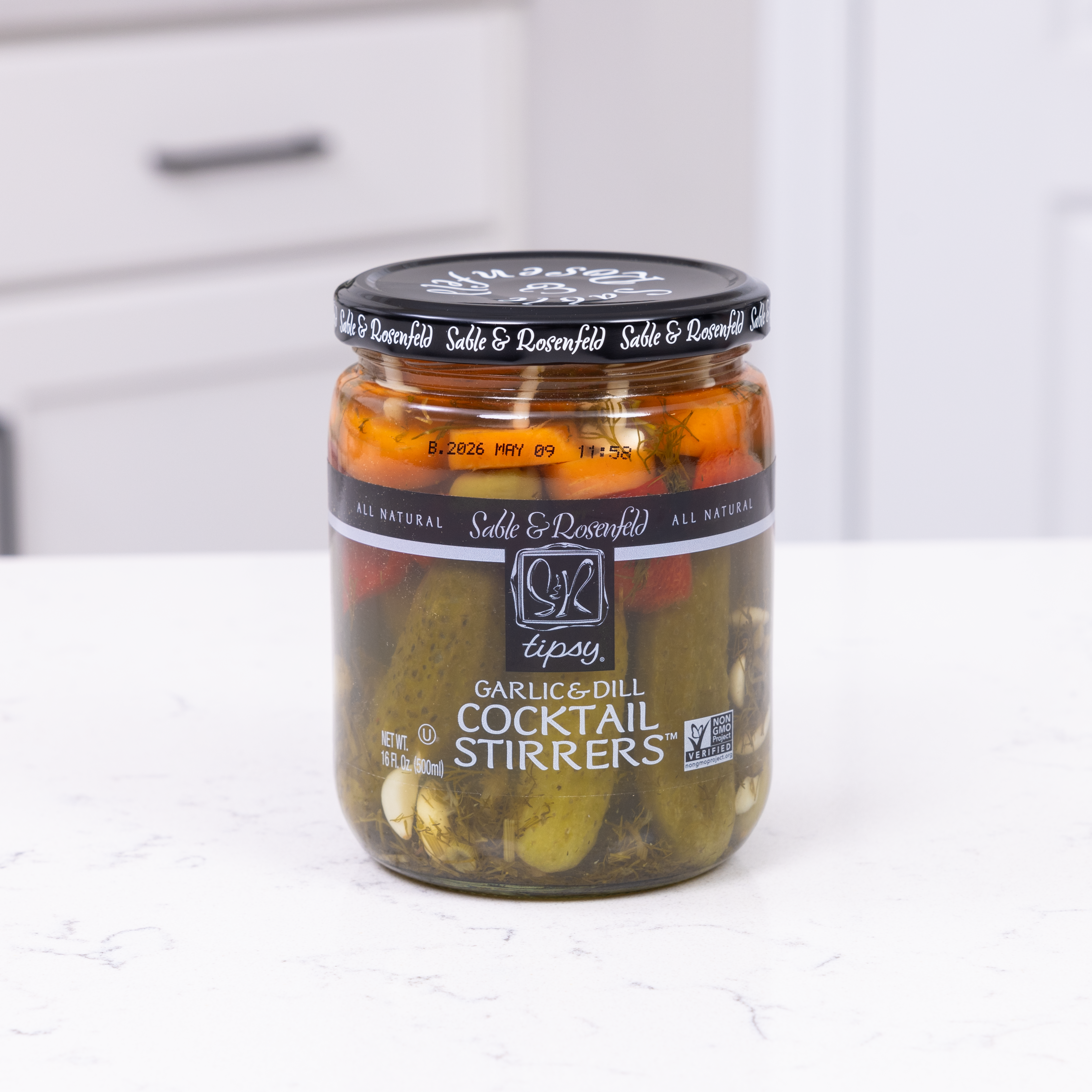 A jar of Sable & Rosenfeld Garlic & Dill Cocktail Stirrers, featuring pickles, peppers, and garlic, placed on a white kitchen countertop—perfect for a cocktail garnish.