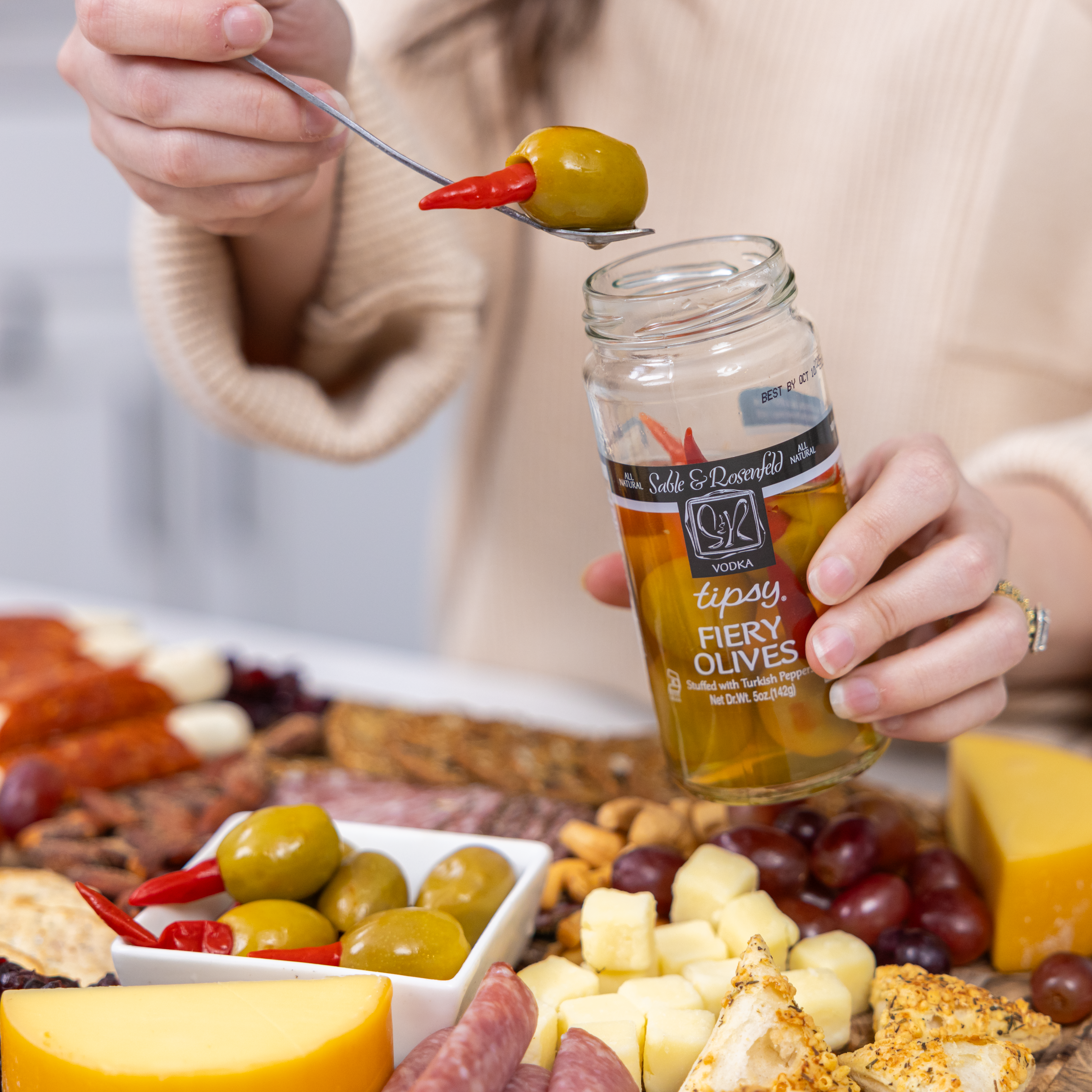 A person holding a jar of Sable & Rosenfeld Tipsy Fiery Olives while picking up an olive stuffed with a Turkish pepper, with a charcuterie board featuring cheese, nuts, grapes, and cured meats in the background.