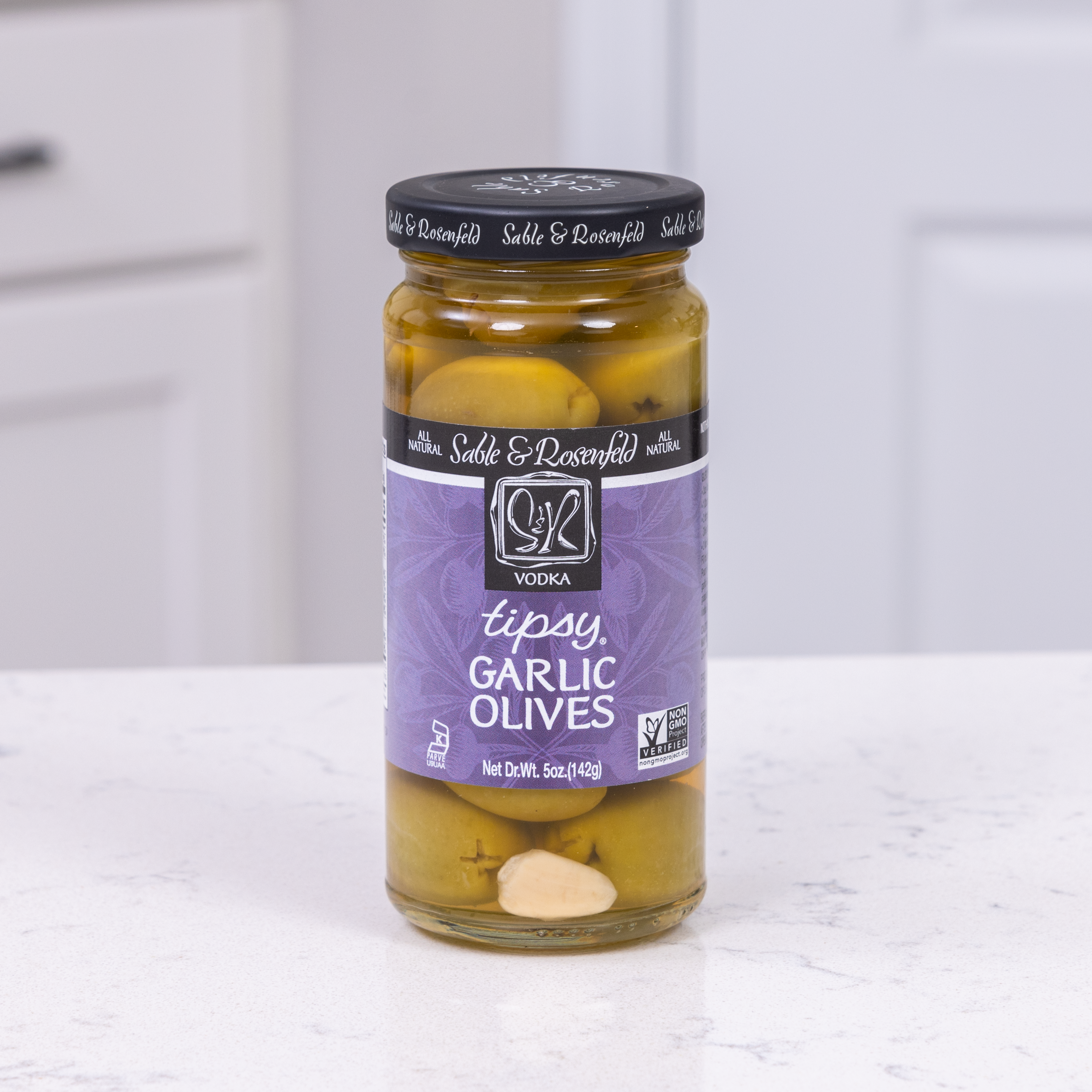 A jar of Sable & Rosenfeld Garlic Stuffed Olives soaked in vodka, placed on a white kitchen countertop.