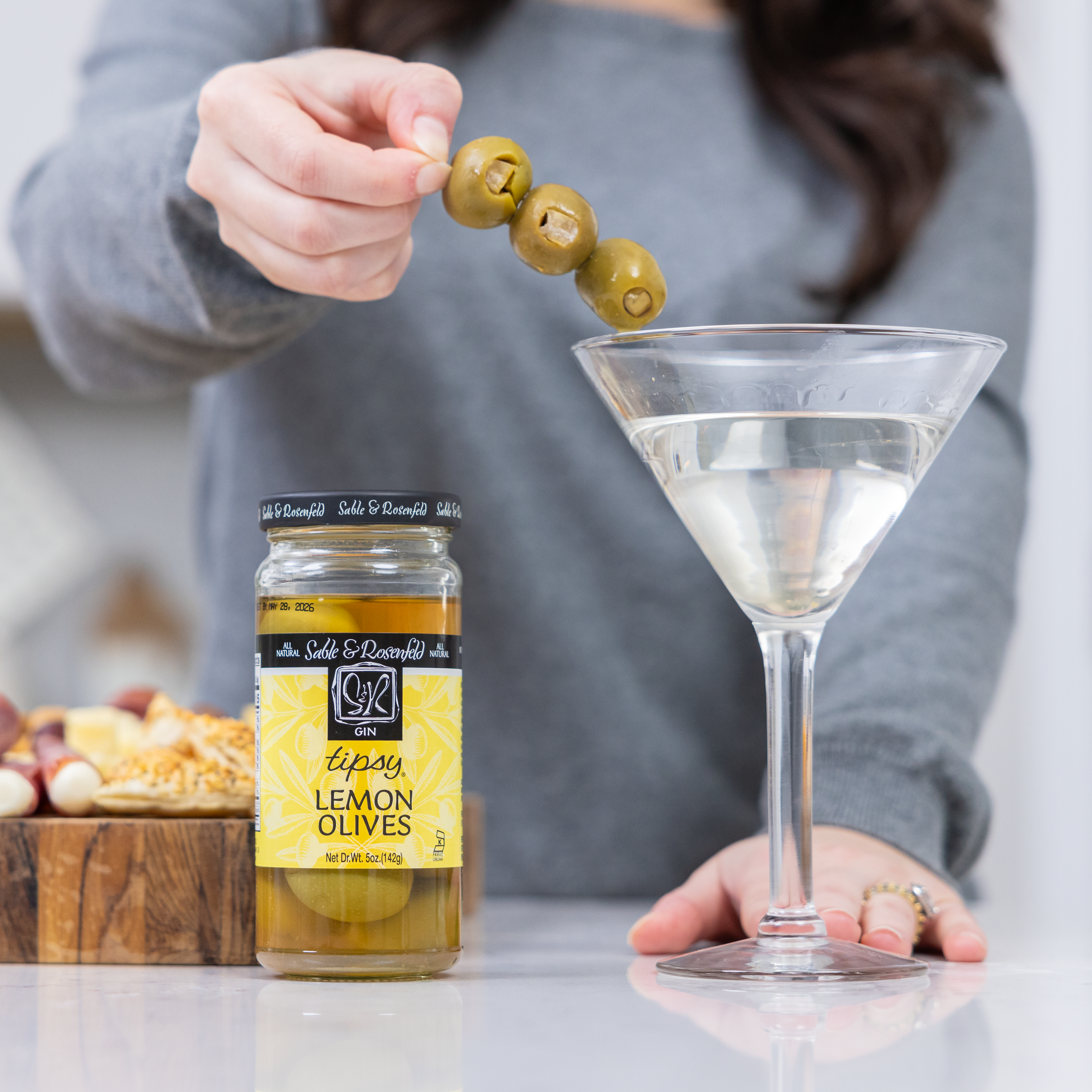 A woman garnishing a gin cocktail with skewered Sable & Rosenfeld Tipsy Lemon Olives, featuring a jar of the vermouth-infused olives on a countertop with a charcuterie board in the background.