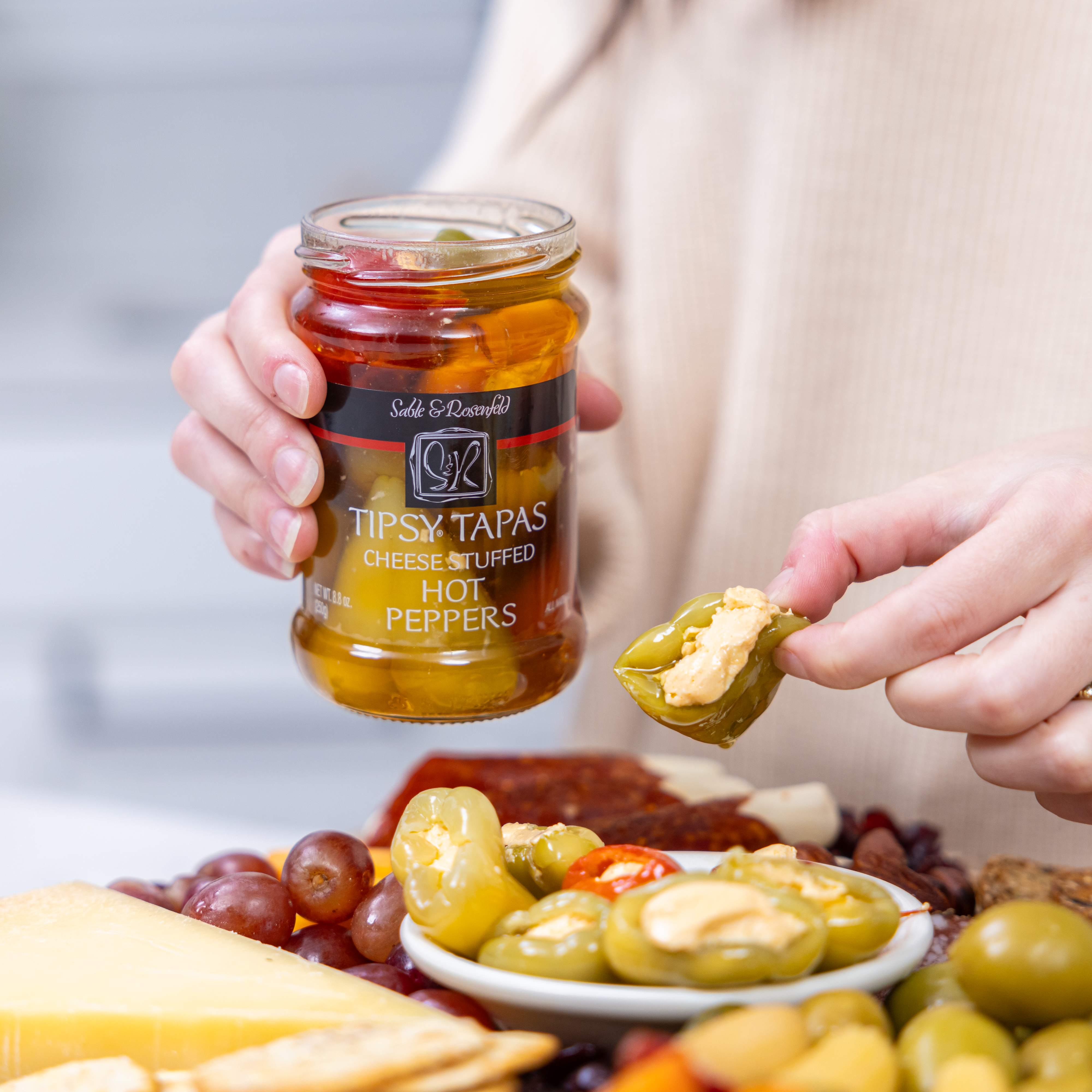 A person holding a jar of Sable & Rosenfeld Tipsy Tapas gourmet stuffed peppers while enjoying a cheese-stuffed hot pepper over a charcuterie board with olives, cheese, and cured meats.