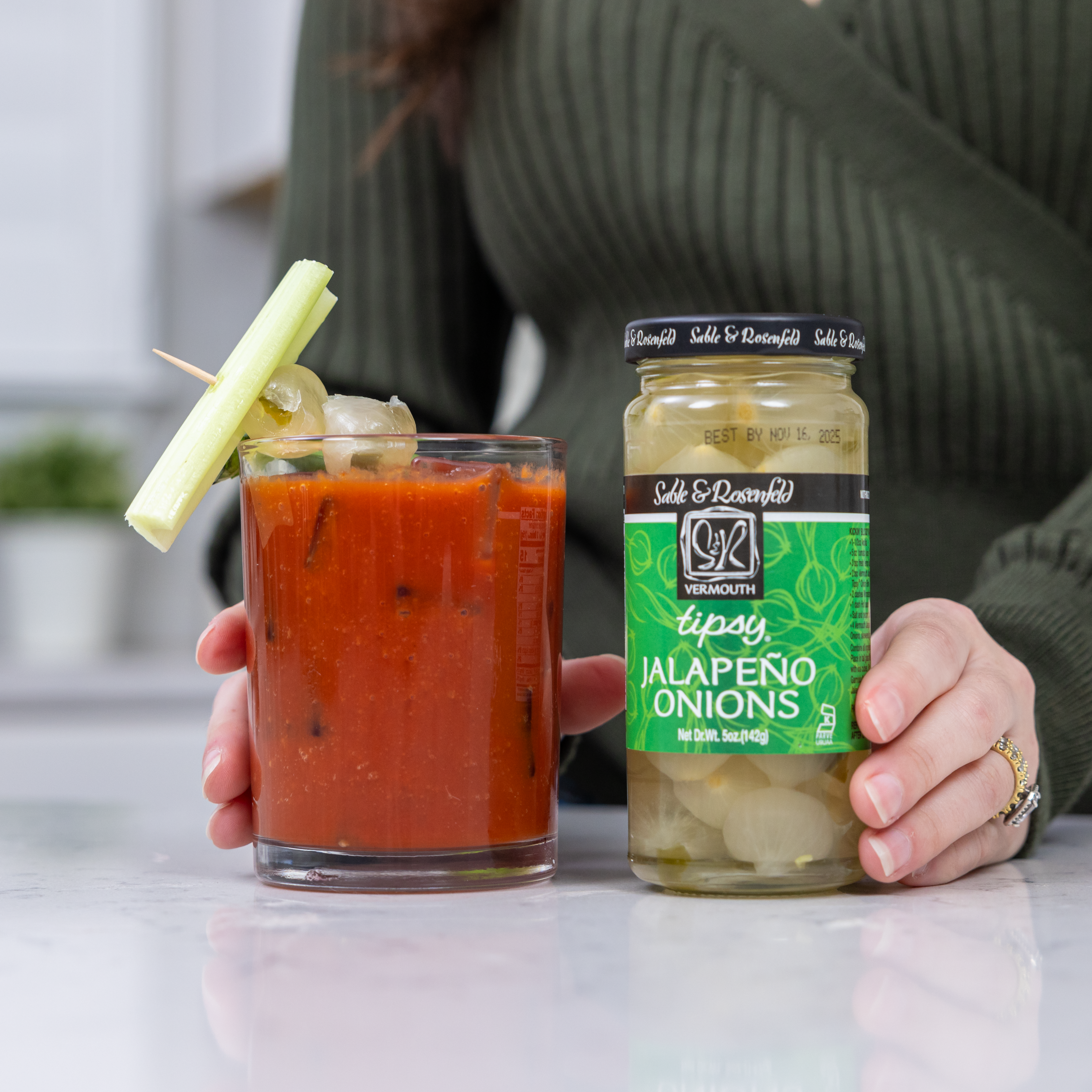 A person holding a Bloody Mary cocktail garnished with celery and Jalapeño Stuffed Onions, next to a jar of Sable & Rosenfeld Tipsy Jalapeño Onions on a white countertop.