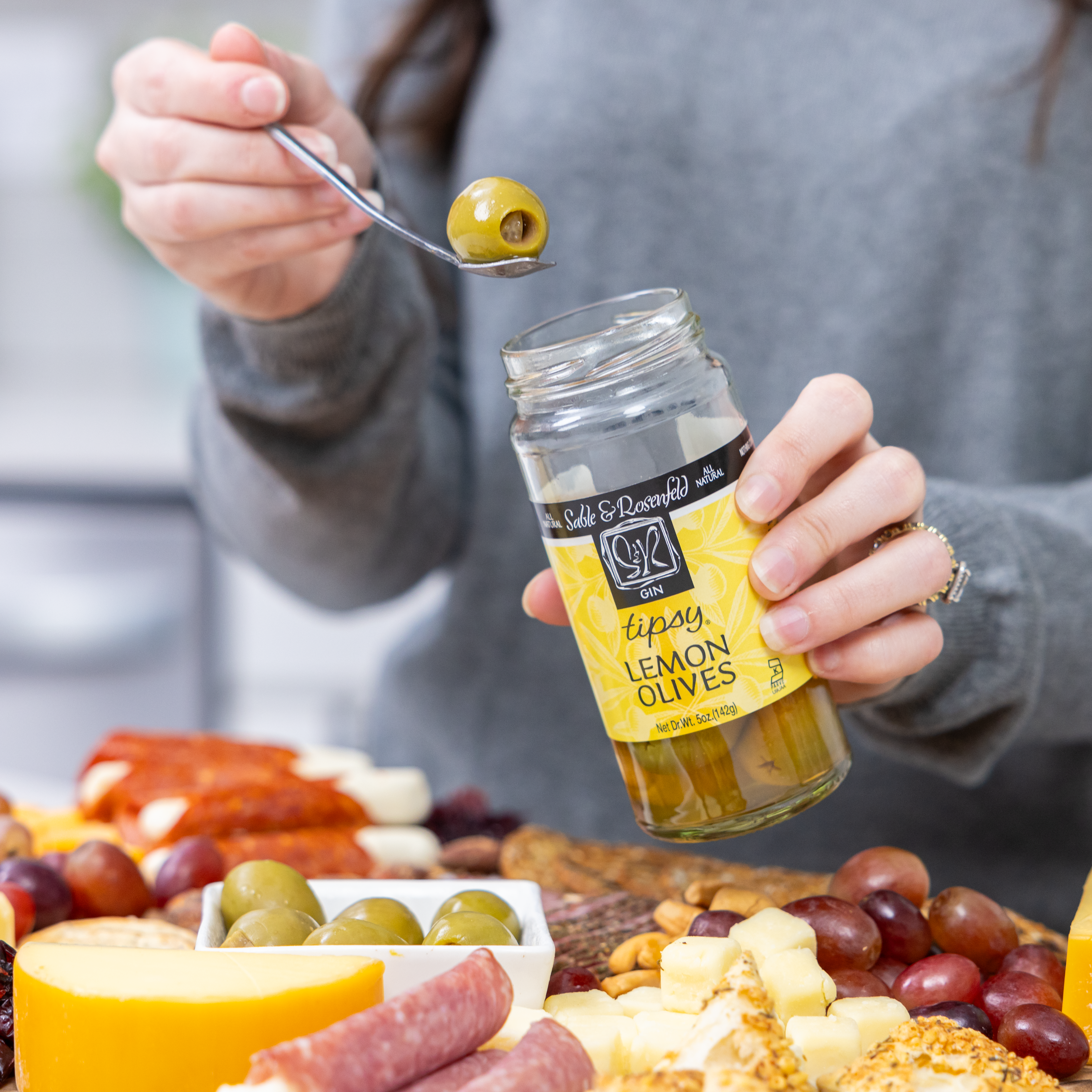 A woman holding a jar of Sable & Rosenfeld Tipsy Lemon Olives while lifting a gin-soaked olive with a spoon over a charcuterie board featuring cheese, crackers, meats, and grapes.