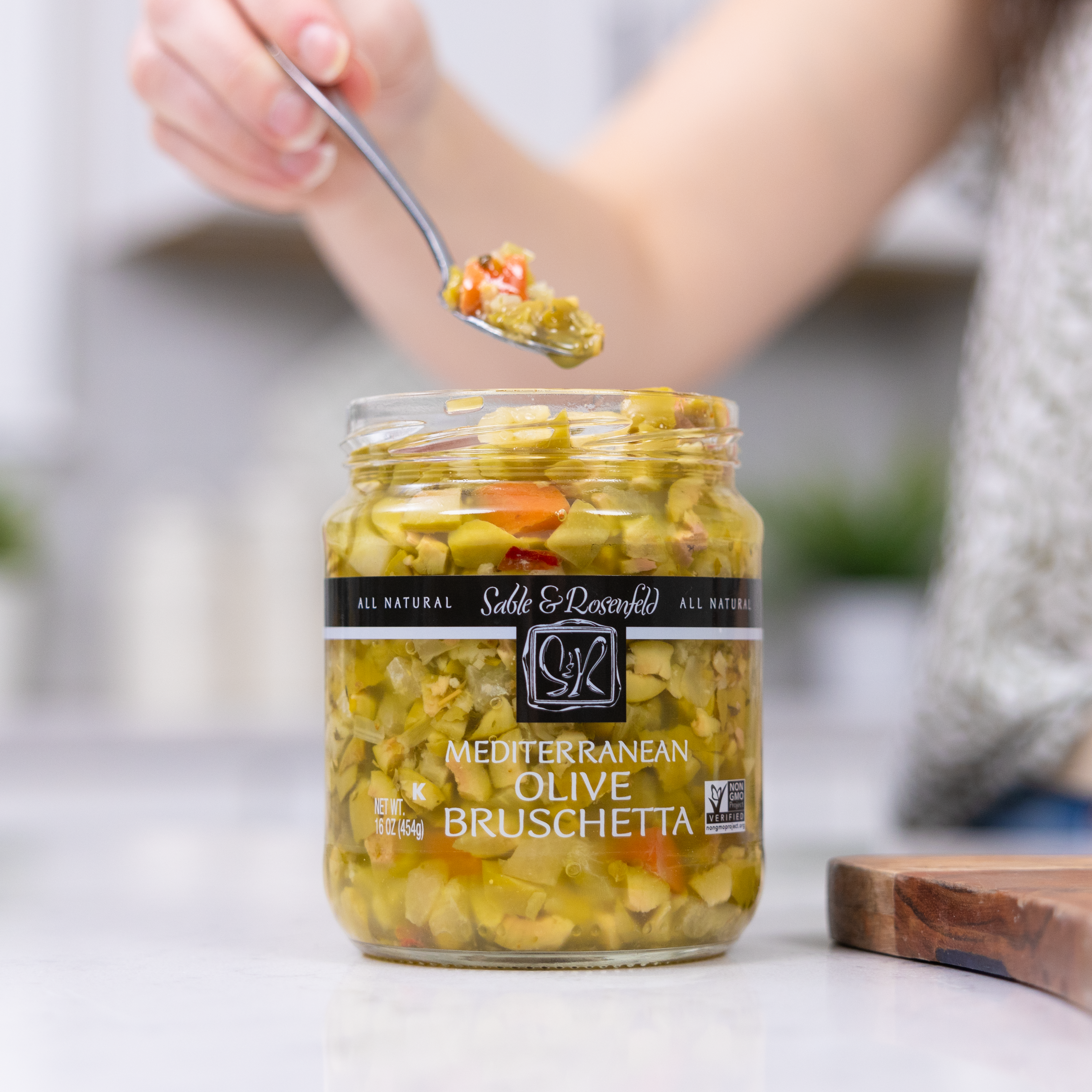 A jar of Sable & Rosenfeld Mediterranean Olive Bruschetta on a kitchen counter, with a person scooping a spoonful of the flavorful olive mixture.