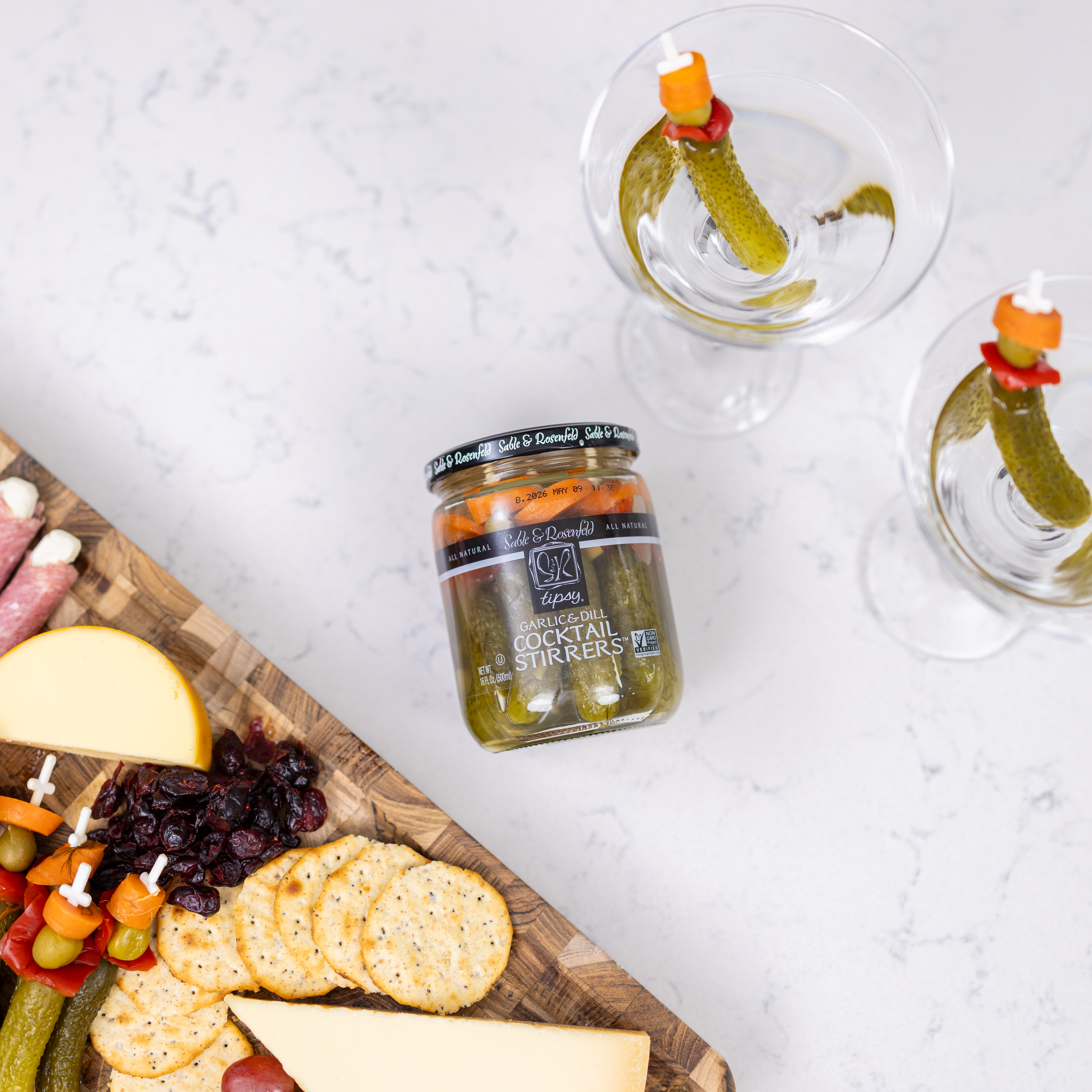 A jar of Sable & Rosenfeld Garlic & Dill Cocktail Stirrers placed next to martini glasses garnished with pickles, peppers, and olives, alongside a charcuterie board— the perfect garnish for cocktails.