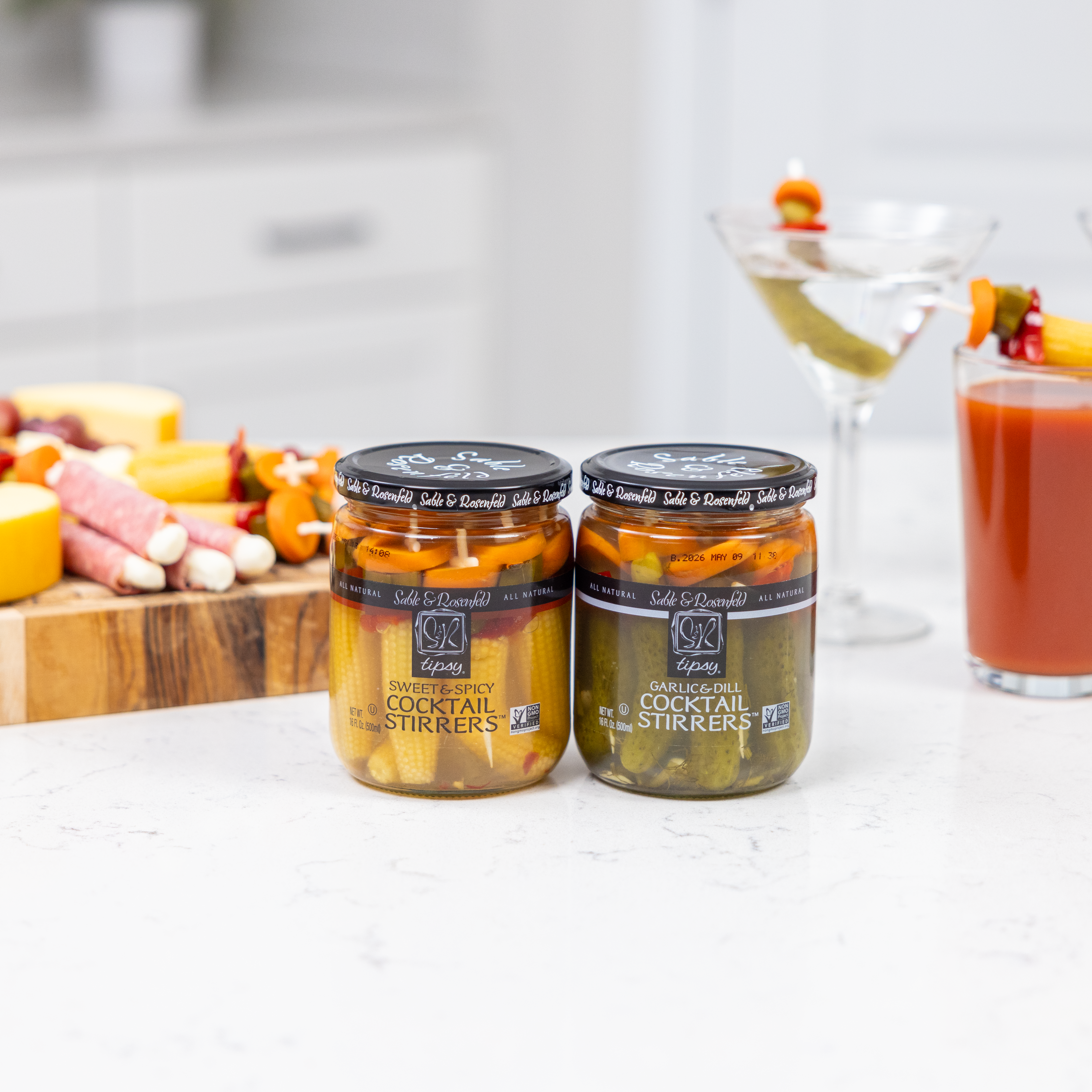 Jars of Sable & Rosenfeld Pickle Cocktail Stirrers in Sweet & Spicy and Garlic & Dill flavors, displayed on a white countertop with martinis and a Bloody Mary in the background.