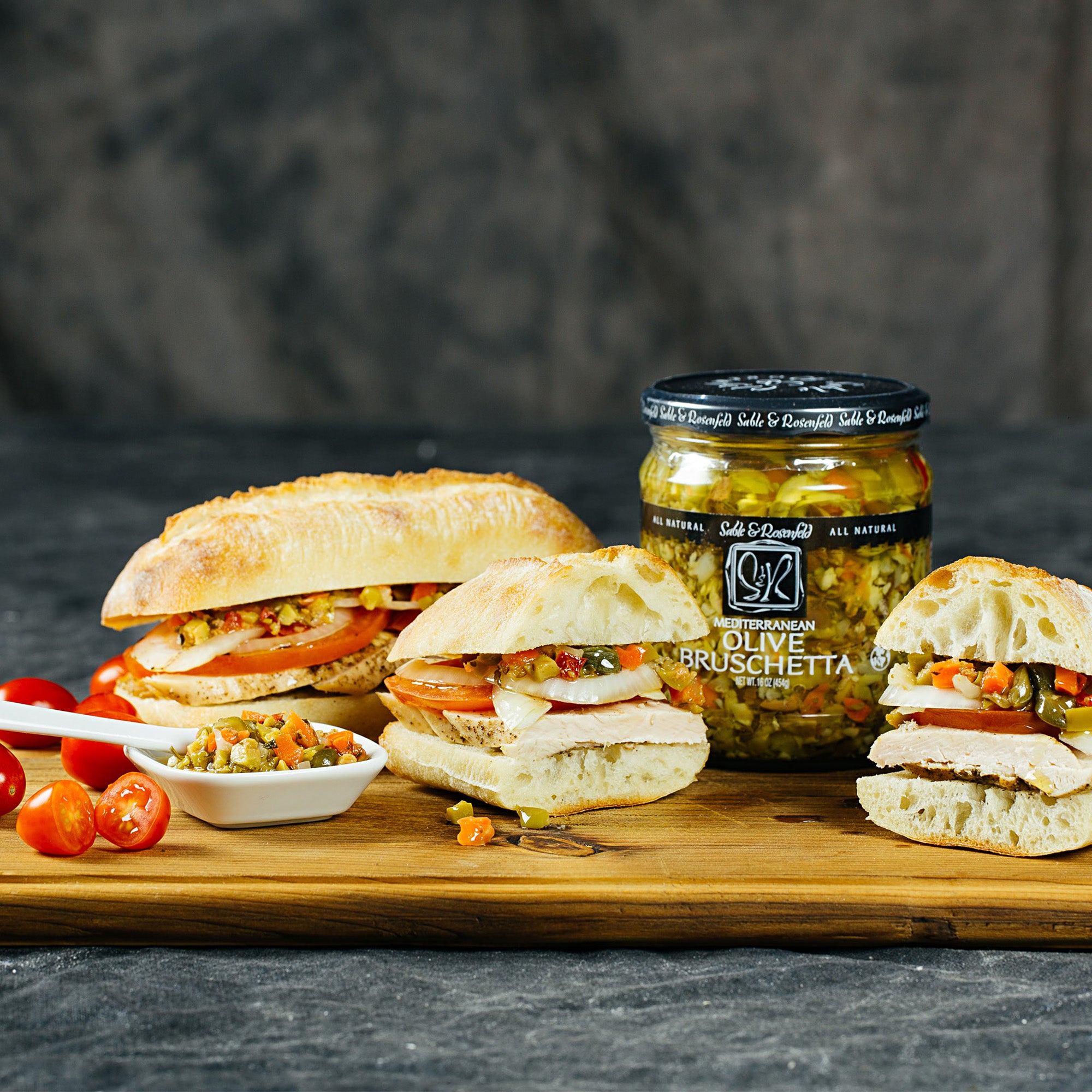 A wooden cutting board displaying an assortment of sandwiches filled with Sable & Rosenfeld Mediterranean Olive Bruschetta, accompanied by fresh tomatoes and a serving dish of the bruschetta mix for a delicious Mediterranean-inspired meal.