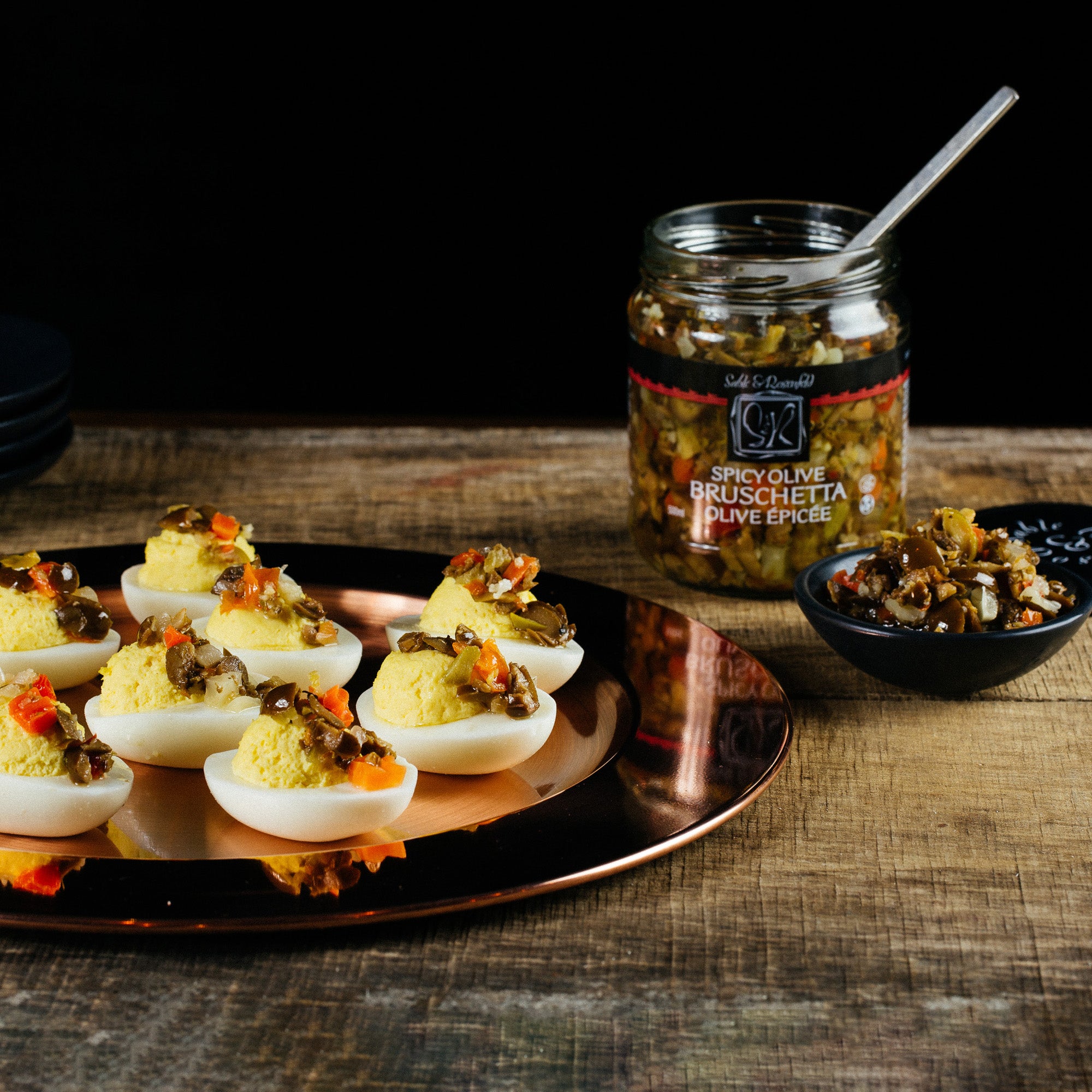 A plate of deviled eggs topped with Sable & Rosenfeld Spicy Olive Bruschetta, served on a rustic wooden table alongside an open jar of the bruschetta and a small bowl filled with the zesty olive mix, creating a delicious and visually appealing appetizer.