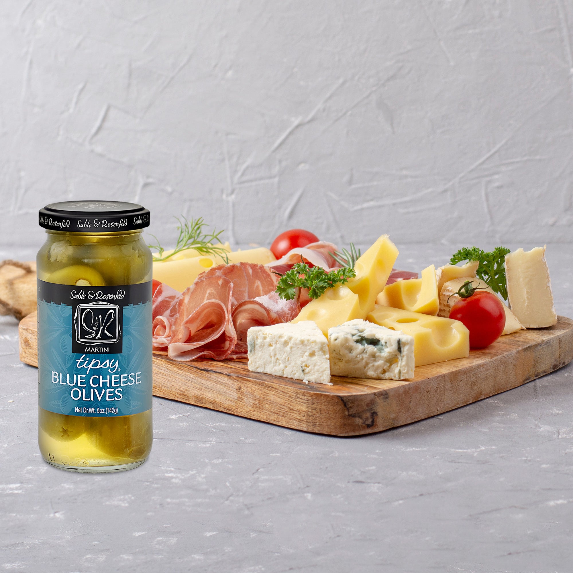 A beautifully arranged cheese platter on a table, featuring a variety of gourmet cheeses, fresh grapes, nuts, and a jar of Sable & Rosenfeld Blue Cheese Tipsy Olives for a flavourful pairing.