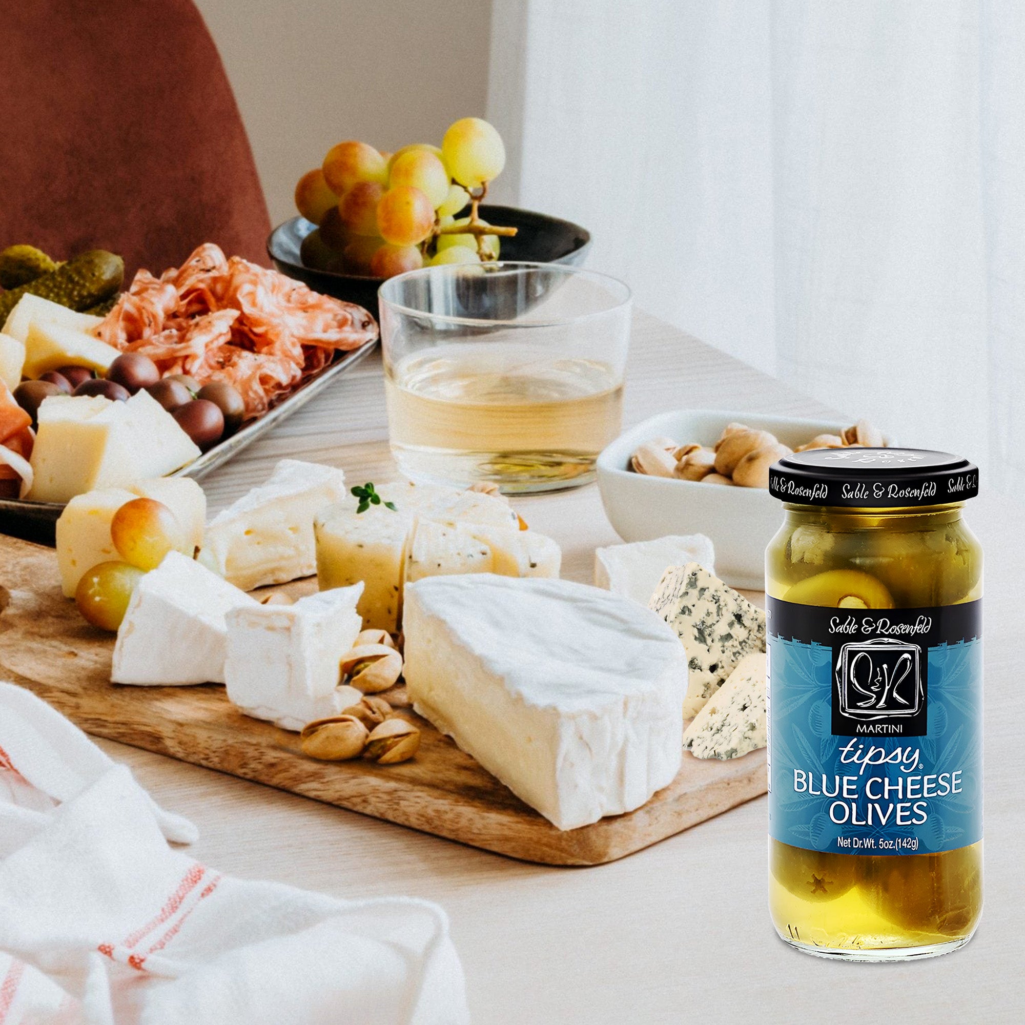 A wooden board set with an assortment of cheeses, meats, and gourmet foods, complemented by a jar of blue cheese-stuffed olives.