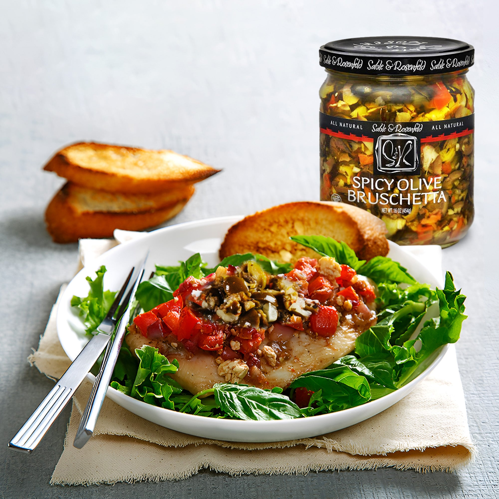 A white plate featuring a fresh salad topped with a generous portion of Sable & Rosenfeld Spicy Olive Bruschetta, served with toasted bread slices for a delicious and vibrant meal presentation.