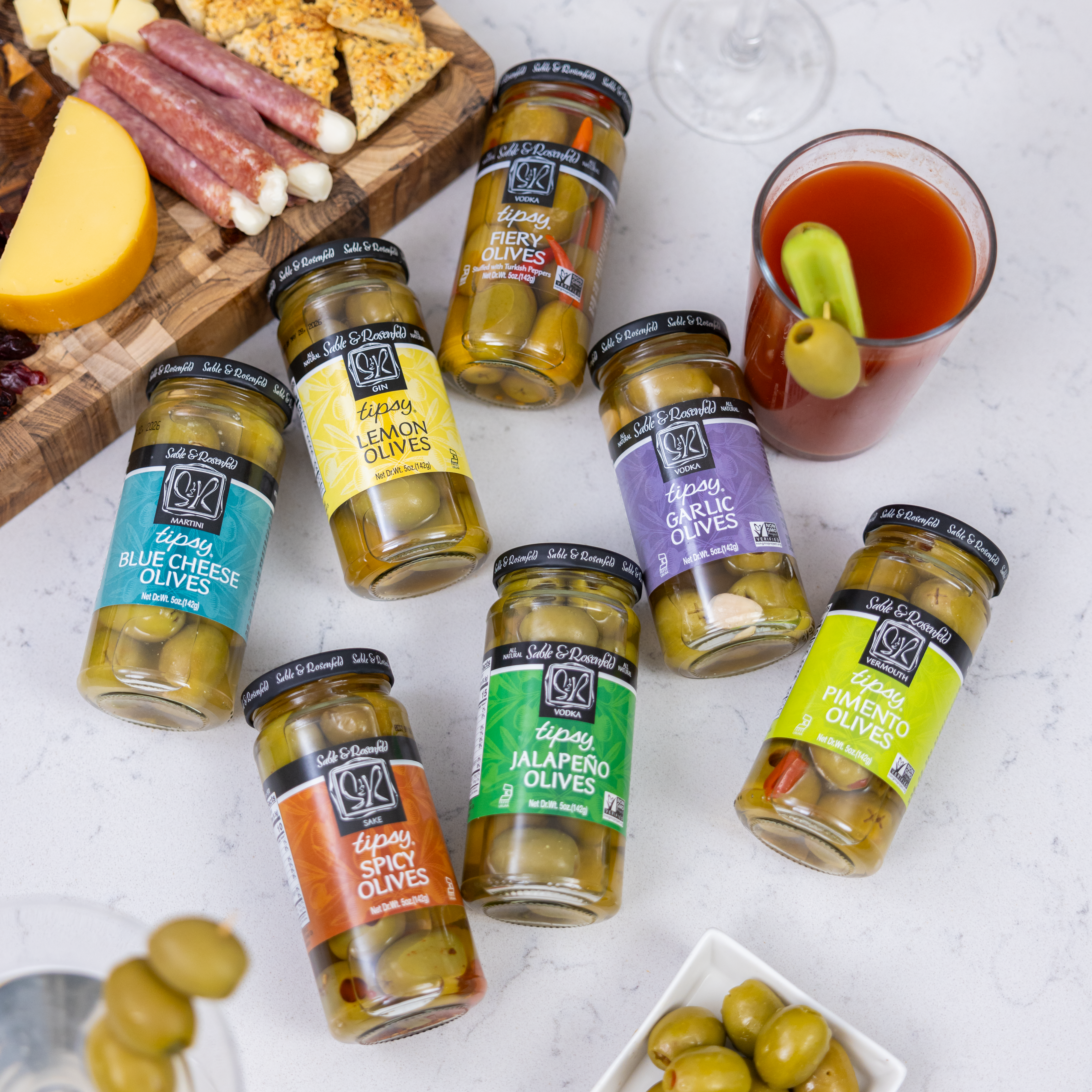 An assortment of Sable & Rosenfeld Tipsy Olives, including Fiery, Lemon, Garlic, Blue Cheese, Pimento, Jalapeño, and Spicy Olives, arranged on a white countertop with a Bloody Mary cocktail and a charcuterie board in the background.