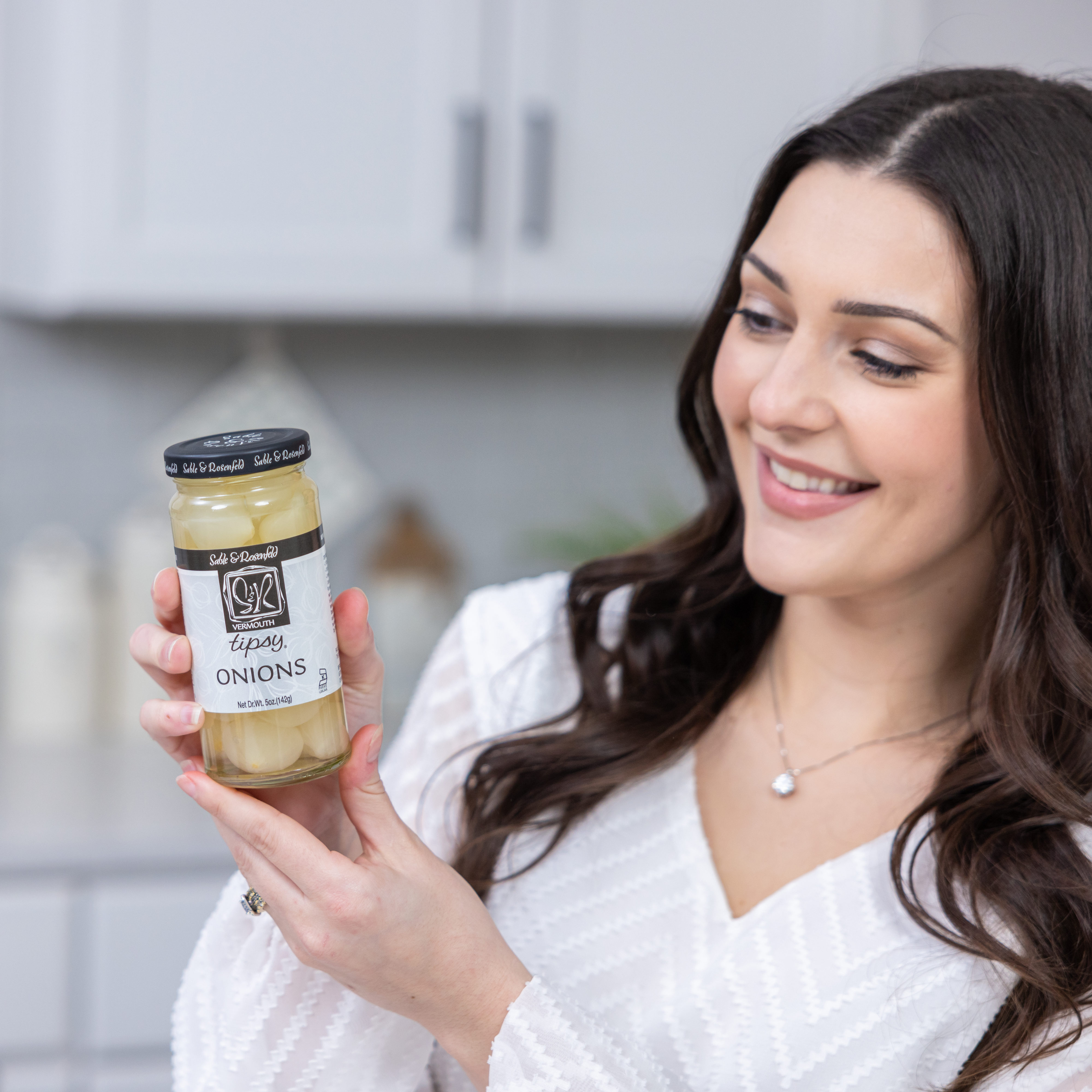 A smiling woman in a white blouse holding a jar of Sable & Rosenfeld Vermouth Tipsy Onions in a modern kitchen. The glass jar contains gourmet cocktail onions infused with dry vermouth, perfect for enhancing martinis and classic cocktails.