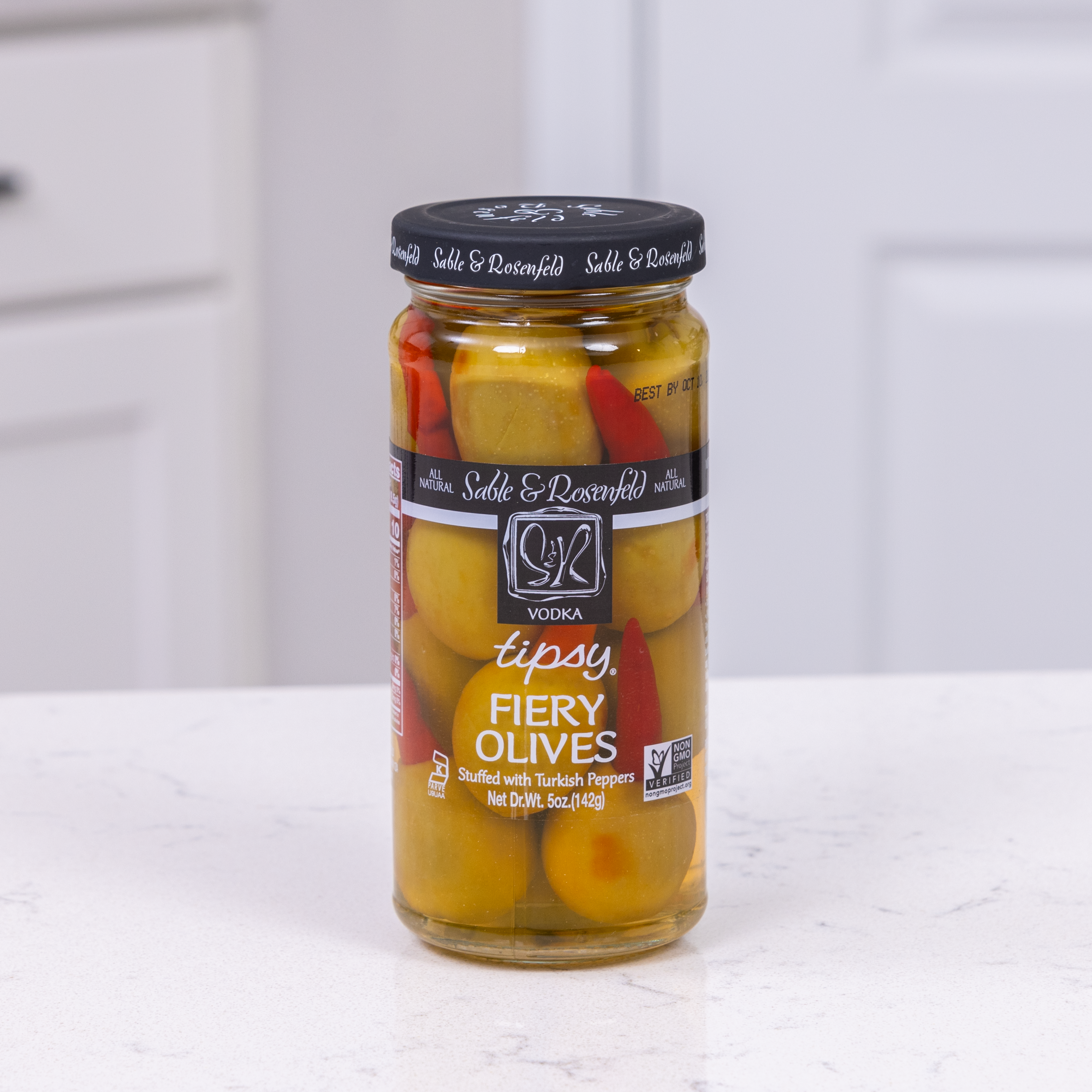 A jar of Sable & Rosenfeld Vodka Tipsy Fiery Olives, featuring green olives stuffed with Turkish peppers, placed on a white kitchen countertop.