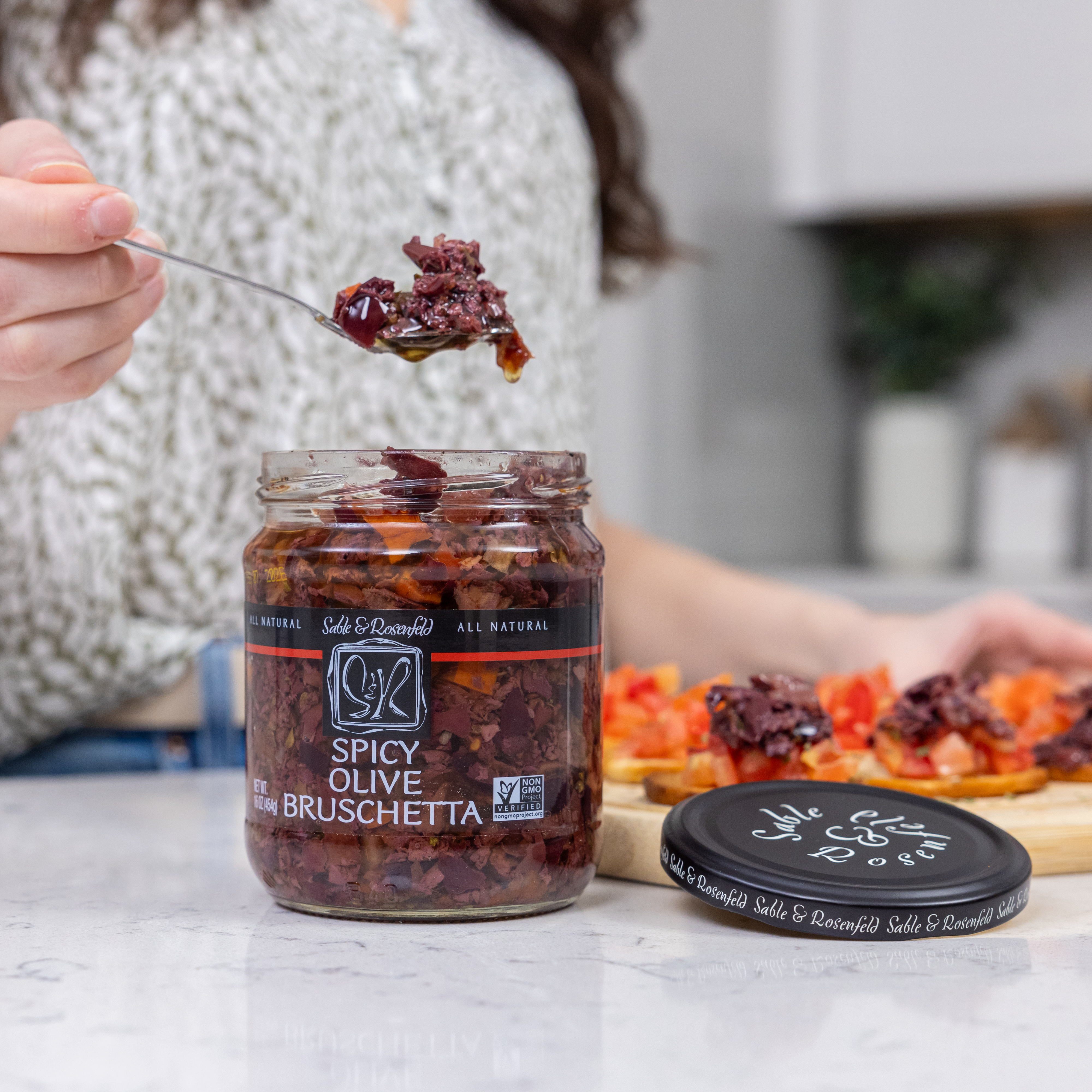 A person scooping a spoonful of Spicy Olive Bruschetta from an open jar, with freshly prepared bruschetta topped with the mixture on a wooden board in the background.