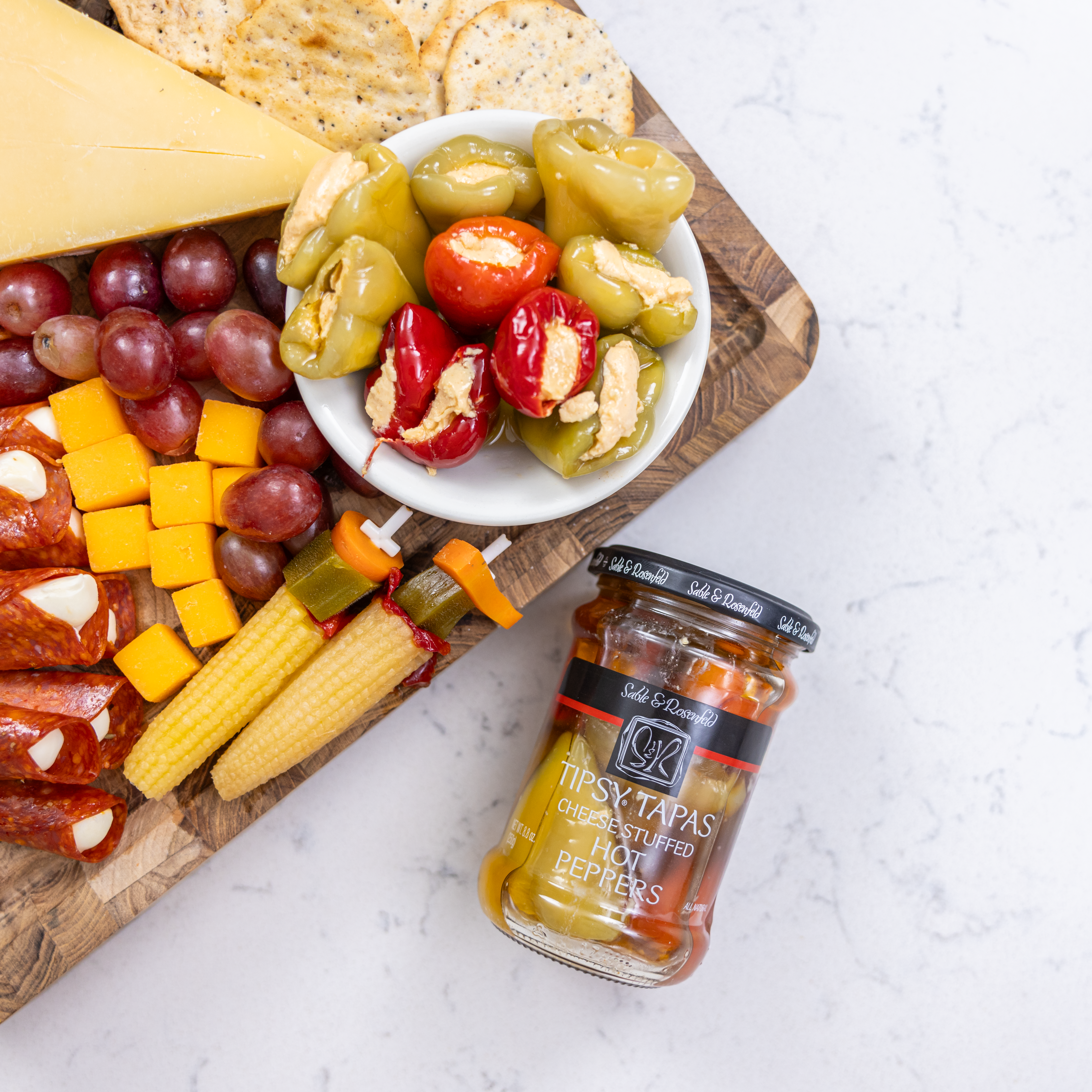 A jar of Sable & Rosenfeld Tipsy Tapas spicy stuffed peppers next to a charcuterie board with a bowl of cheese-stuffed hot peppers, grapes, cheese cubes, crackers, and cured meats.