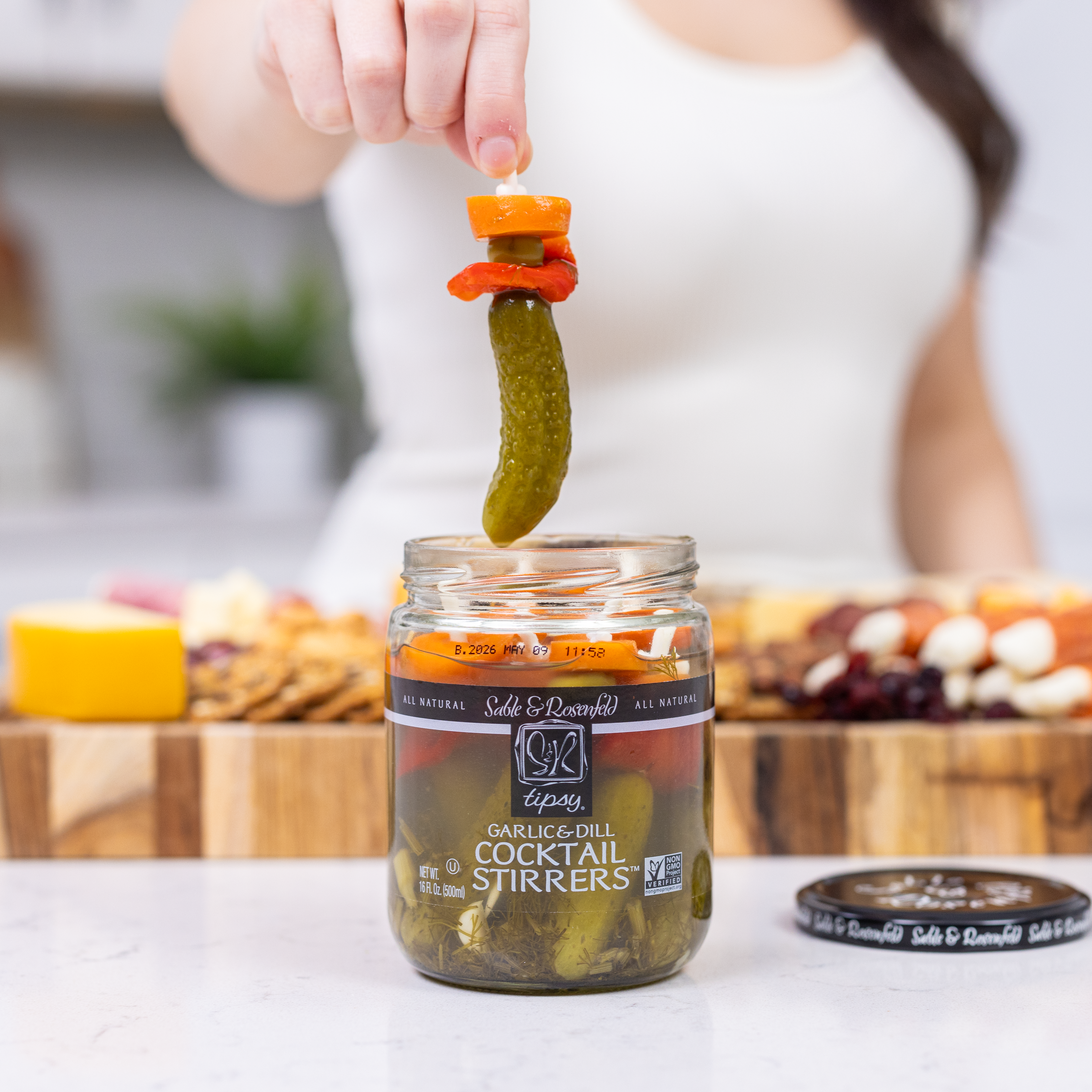 A jar of Sable & Rosenfeld Tipsy Garlic & Dill Cocktail Stirrers, featuring pickled gherkins and vegetables, with a person lifting a skewer from the jar against a gourmet charcuterie board background.