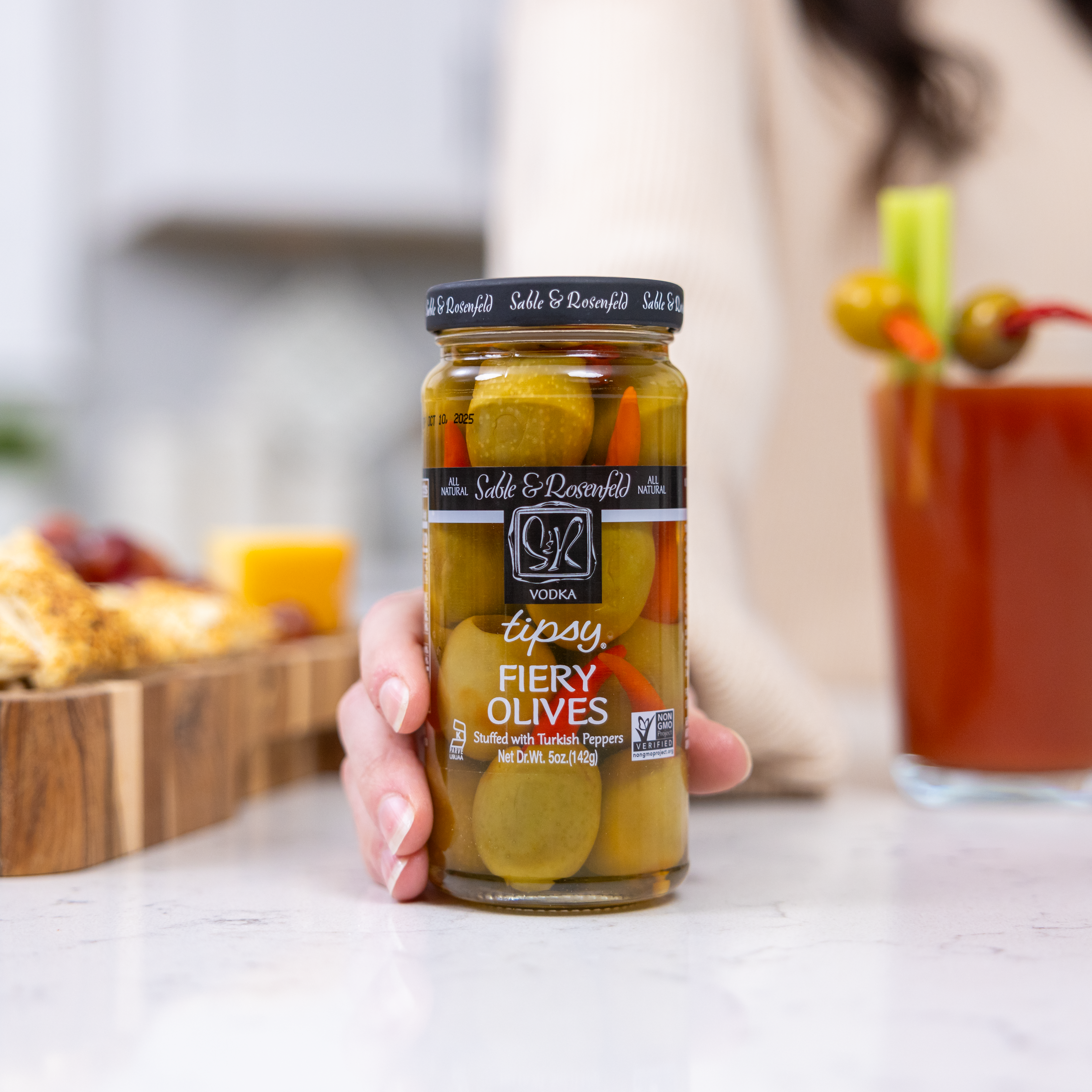 A hand holding a jar of Tipsy Fiery Olives by Sable & Rosenfeld, infused with vodka and stuffed with Turkish peppers, with a Bloody Mary cocktail and appetizers in the background.