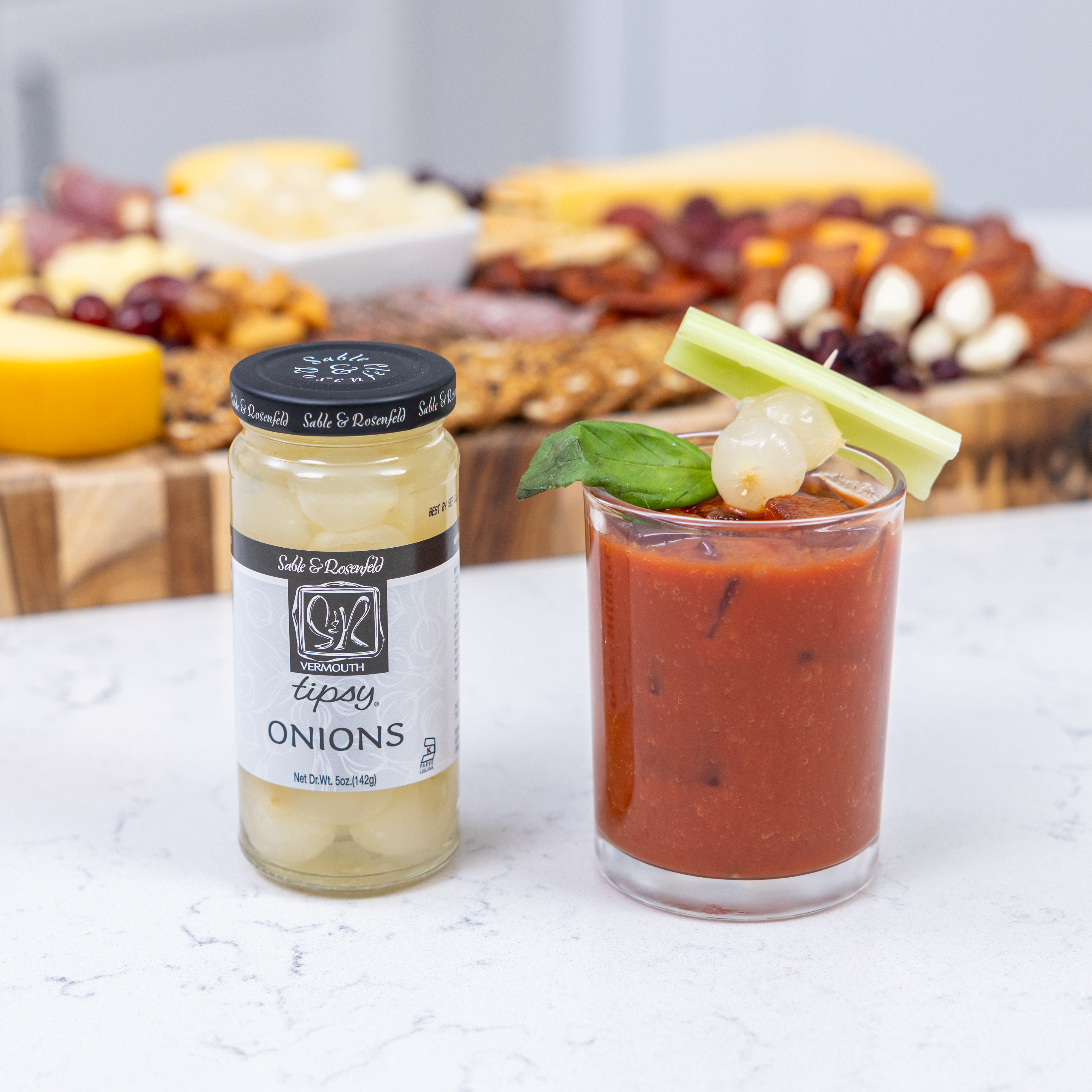 A jar of Vermouth-infused pickled onions next to a Bloody Mary cocktail, garnished with a celery stick, basil leaf, and cocktail pick holding pickled onions. A gourmet charcuterie board with cheeses and meats is visible in the background.