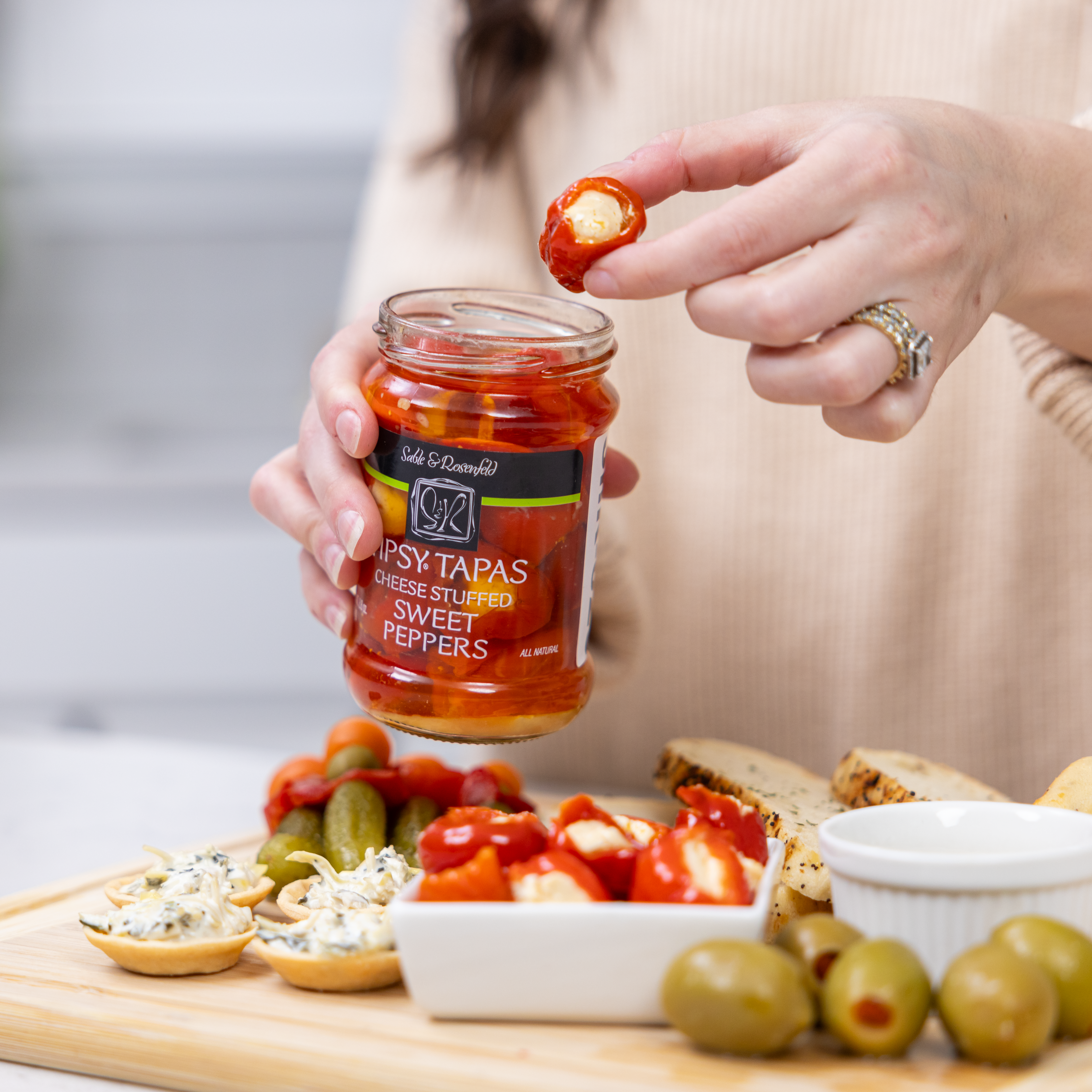 Jar of Tipsy Tapas cheese stuffed sweet peppers held by a woman, with a gourmet appetizer platter featuring olives, crackers, and stuffed peppers.