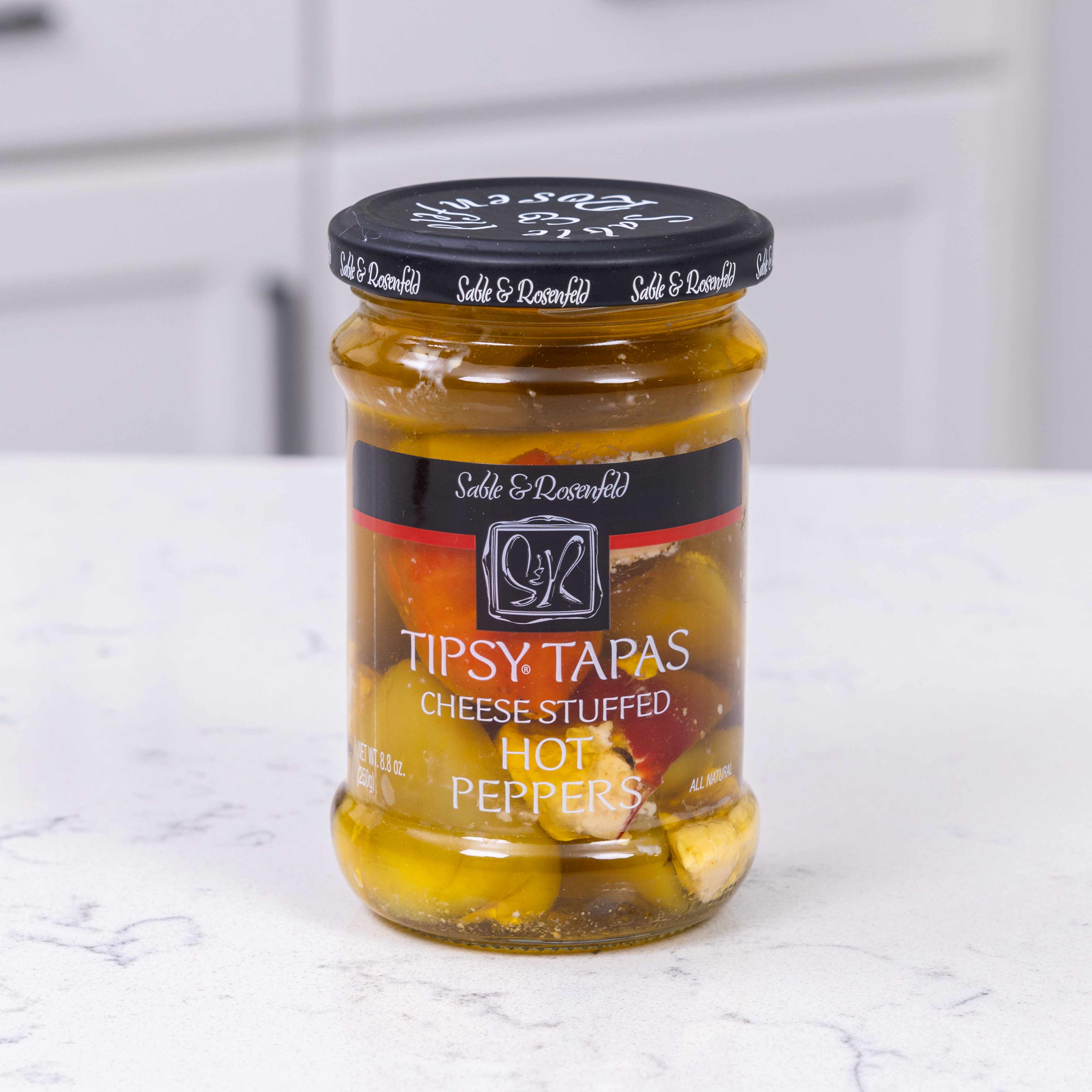 A jar of Sable & Rosenfeld Tipsy Tapas Hot Peppers, featuring cheese-stuffed red and yellow peppers in brine, placed on a white kitchen countertop.