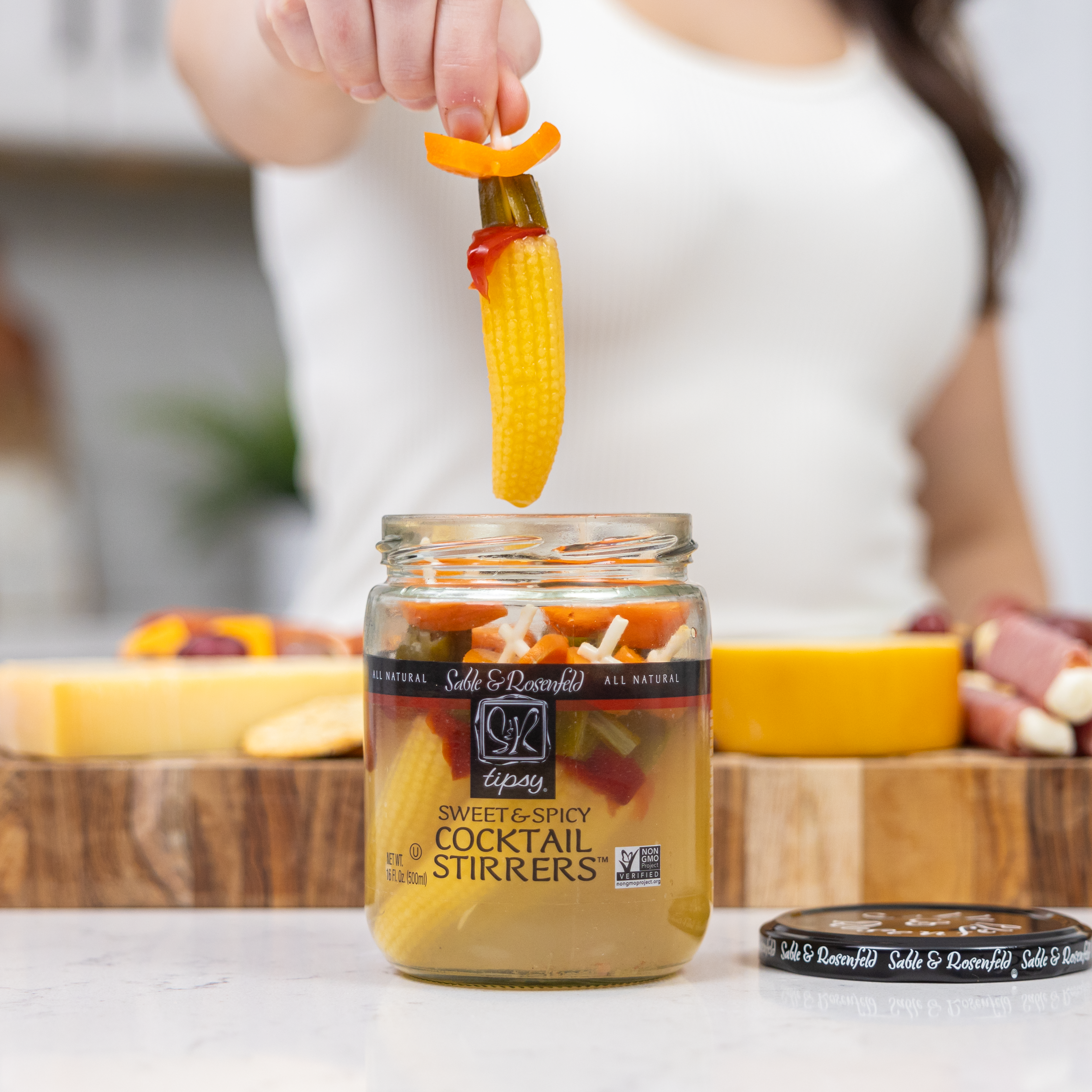 A person holding a Sable & Rosenfeld Tipsy Cocktail Stirrer, featuring baby corn, pickles, and peppers, above an open jar of Sweet & Spicy Cocktail Stirrers on a kitchen countertop.