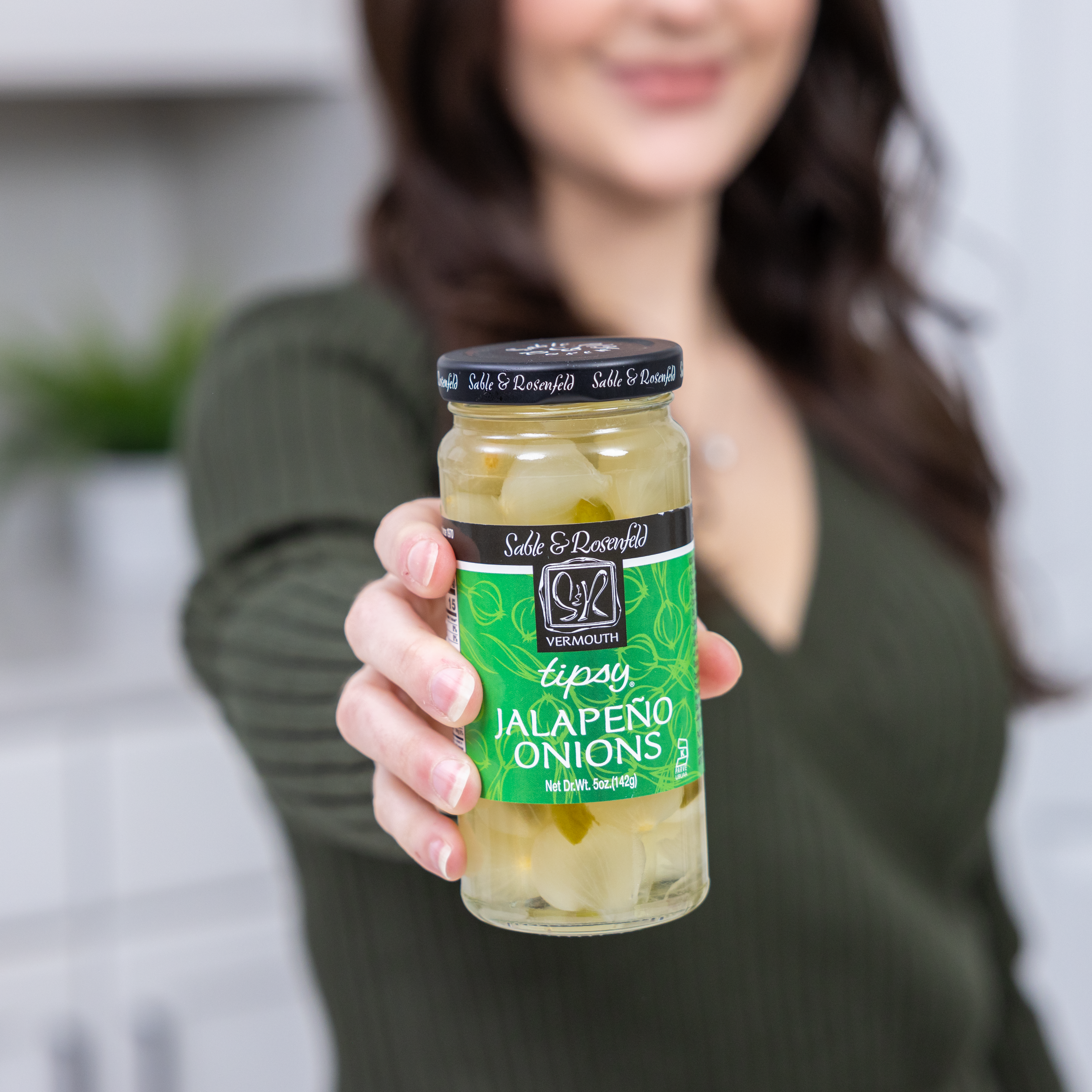 A woman holding a jar of Sable & Rosenfeld Vermouth Jalapeño Tipsy Onions, featuring cocktail onions infused with vermouth and jalapeño, against a blurred kitchen background.