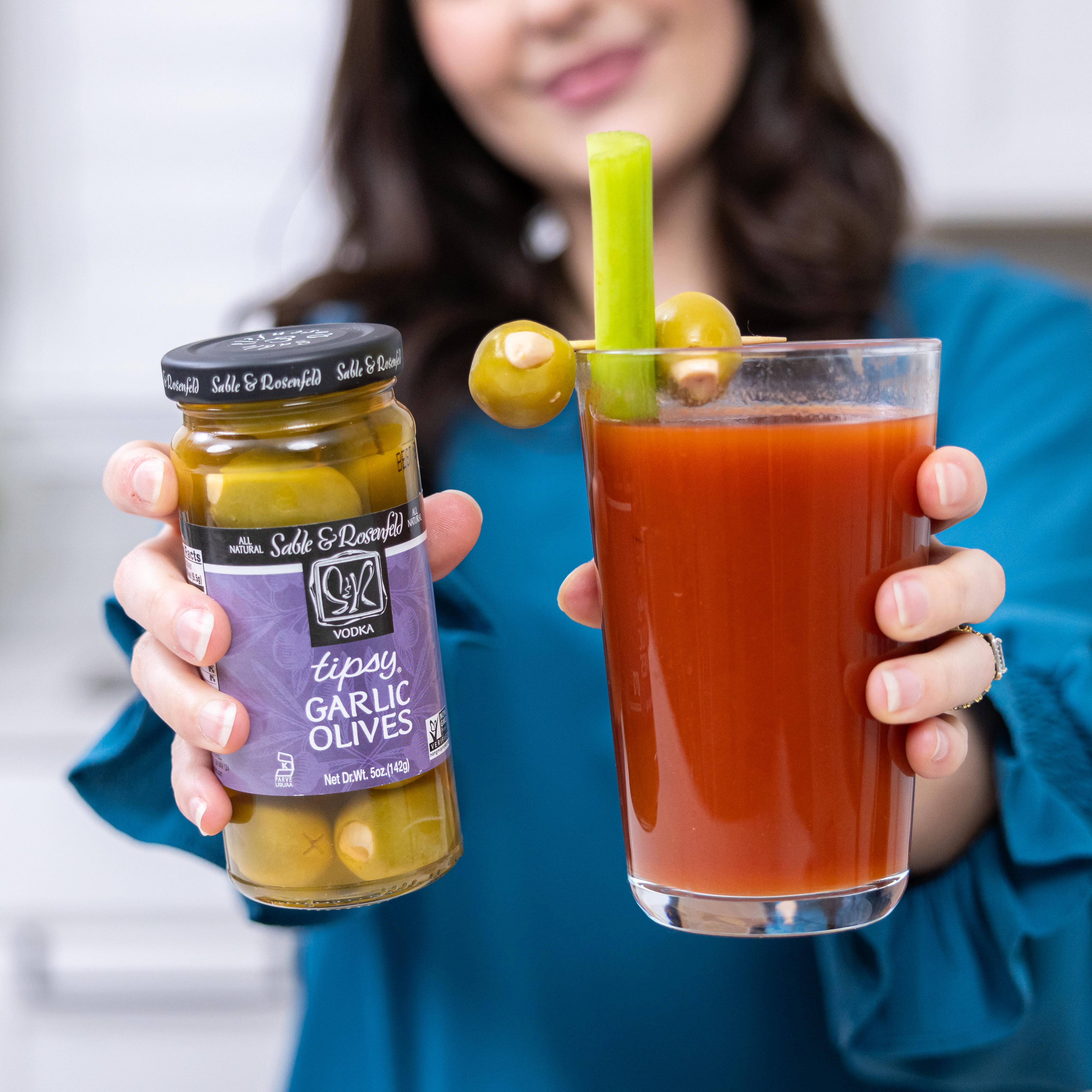 A woman holding a jar of Sable & Rosenfeld Vodka Garlic Tipsy Olives in one hand and a Bloody Mary garnished with garlic-stuffed olives and a celery stick in the other.