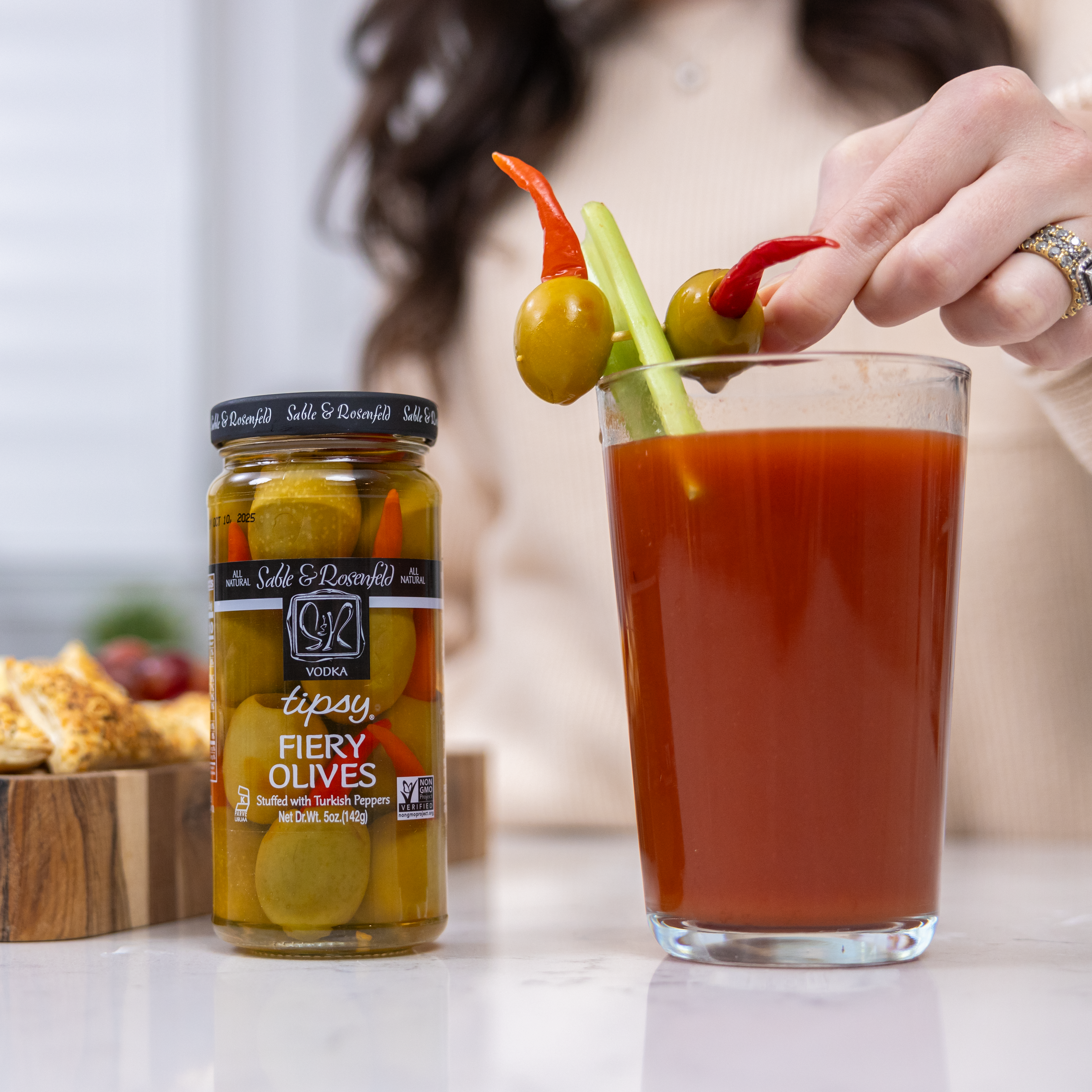 A person garnishing a Bloody Mary with Vodka Tipsy Fiery Olives by Sable & Rosenfeld, featuring green olives stuffed with Turkish peppers on a celery stick. A jar of Vodka Tipsy Fiery Olives is placed beside the cocktail on a white countertop.