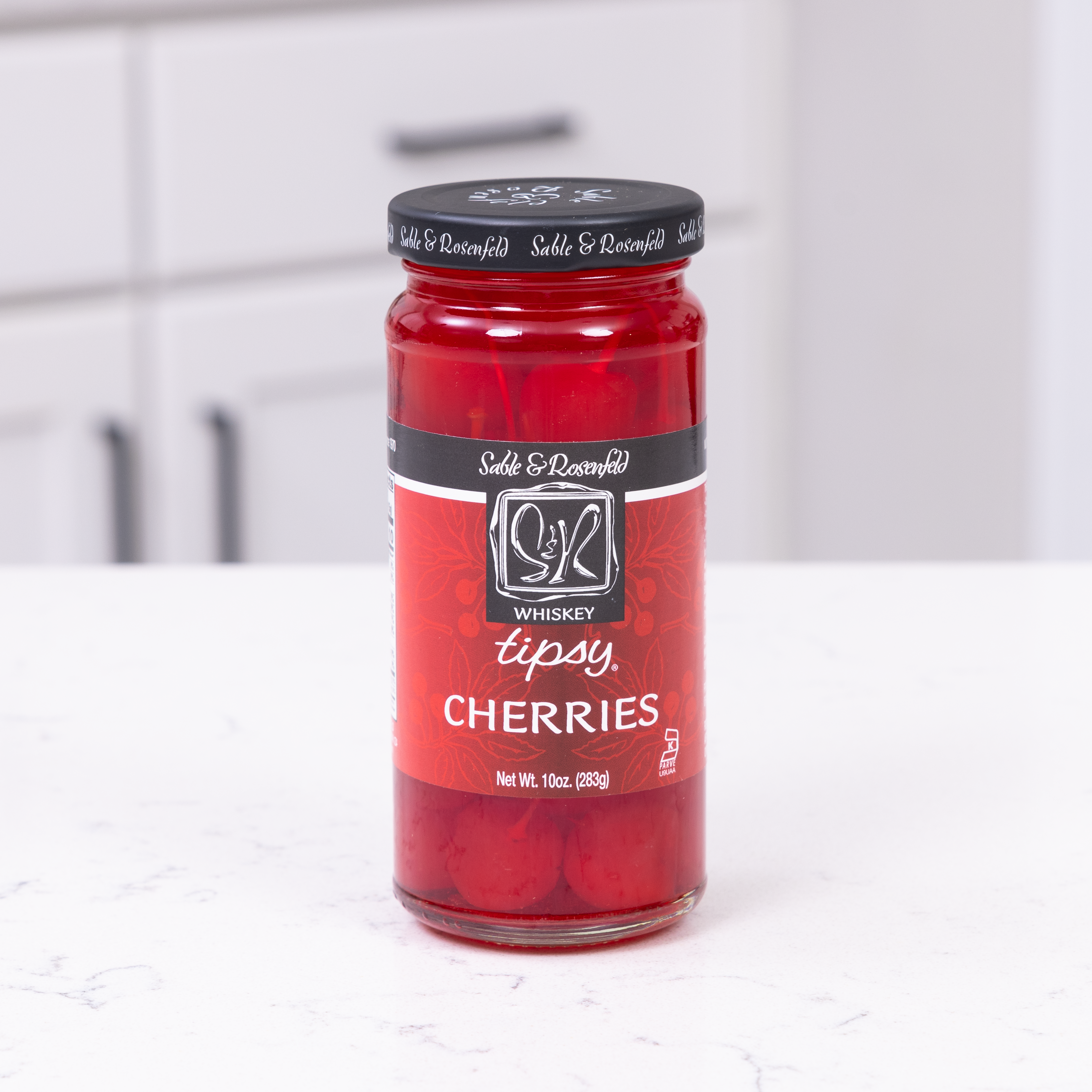 A jar of Sable & Rosenfeld Whiskey Tipsy Cherries placed on a white marble countertop in a modern kitchen. The glass jar showcases vibrant red cherries soaked in whiskey, perfect for cocktails and gourmet desserts.