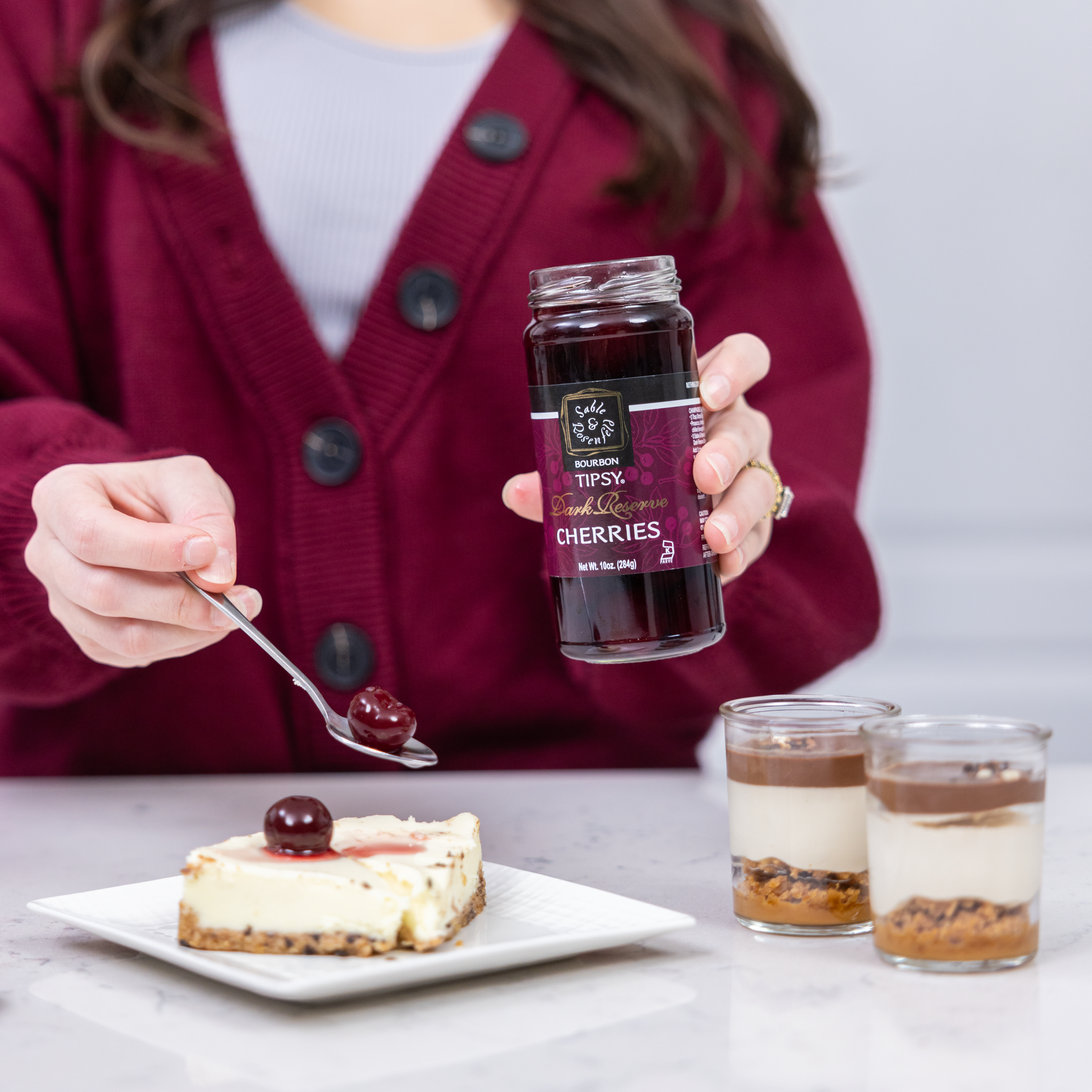 Woman holding a jar of black cherry bourbon infused coffee cherries, garnishing a cheesecake dessert.