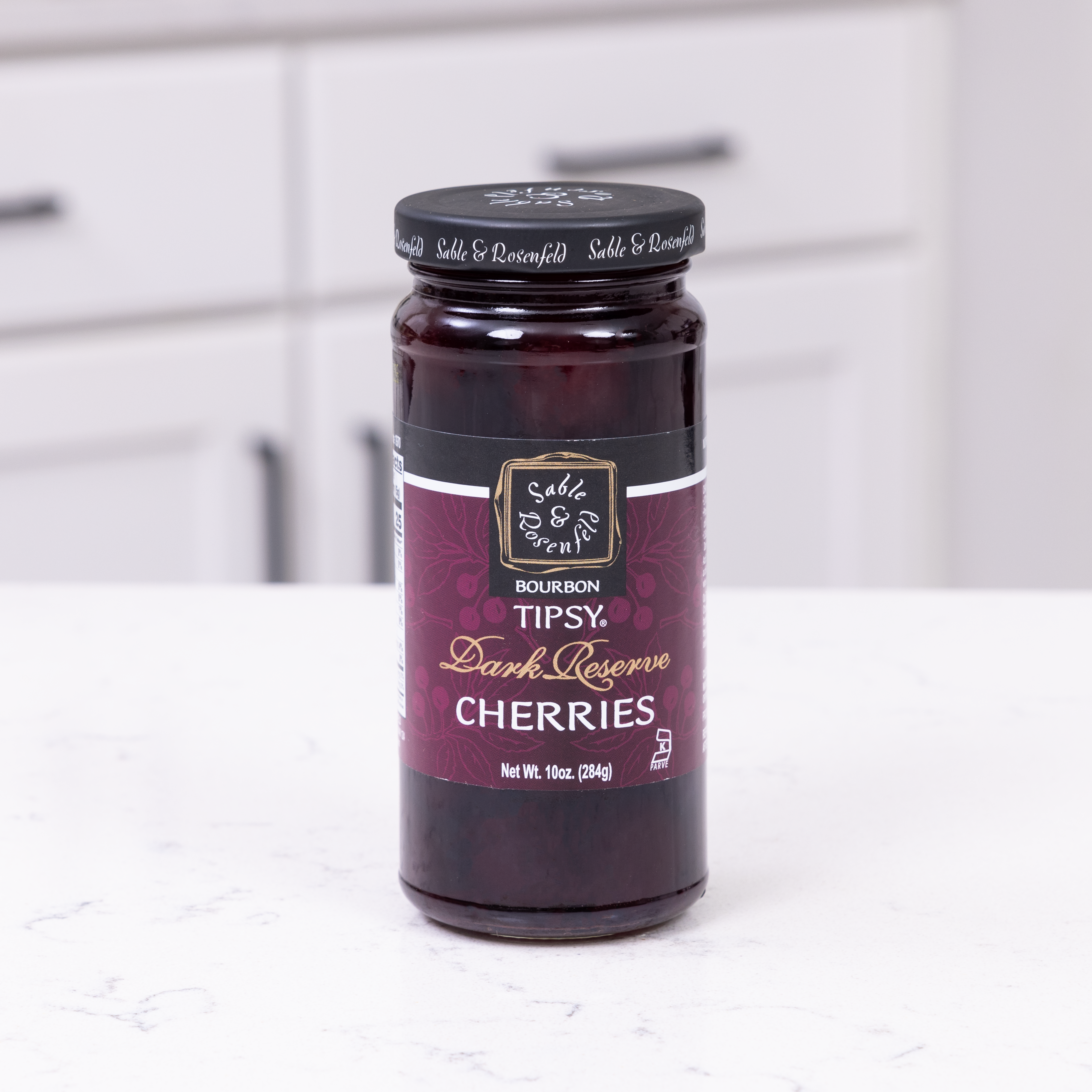 Jar of black cherry infused bourbon cherries on a white kitchen counter, perfect for cocktails and desserts.