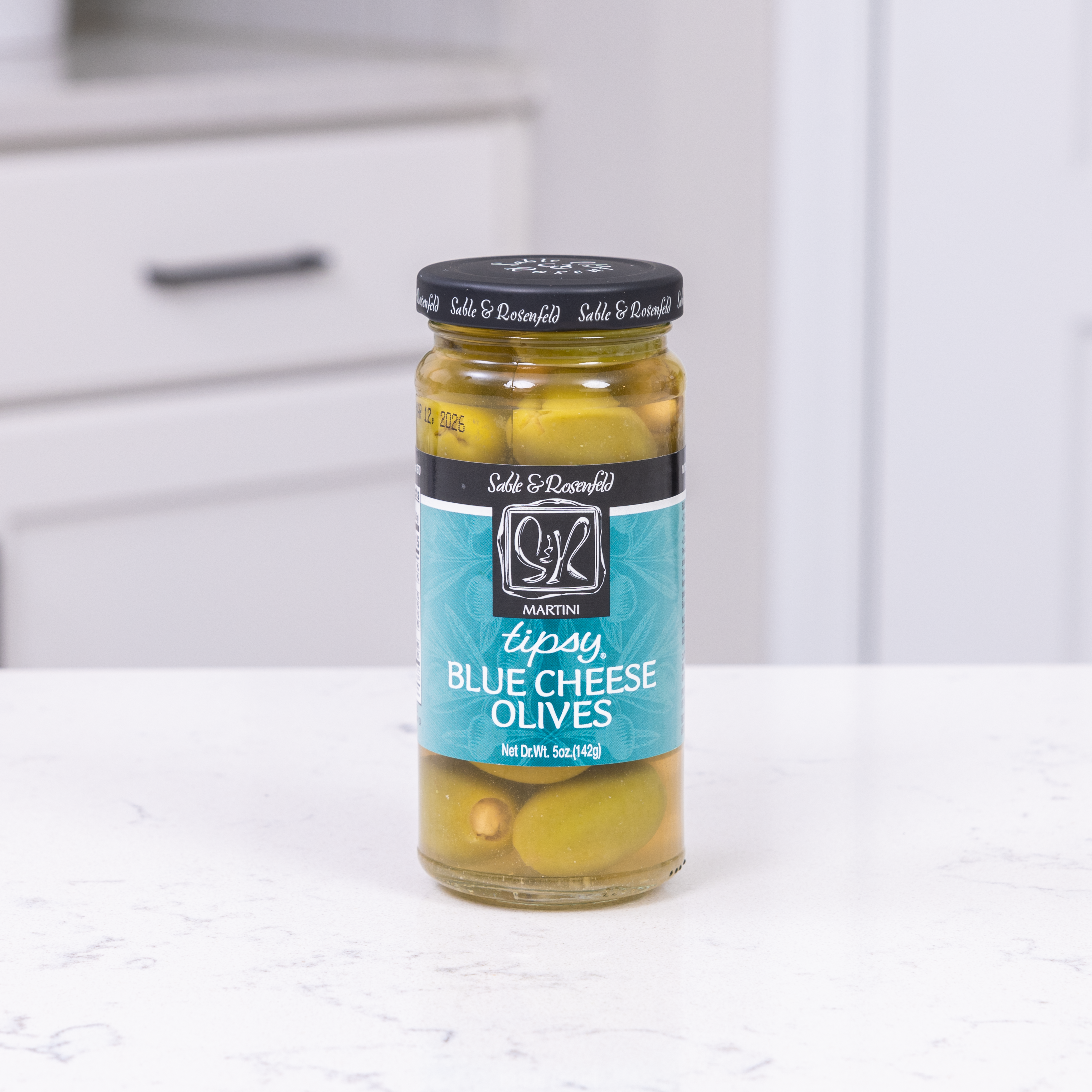 A jar of Sable & Rosenfeld Tipsy Blue Cheese Olives placed on a white kitchen countertop, showcasing delicious green olives filled with blue cheese.