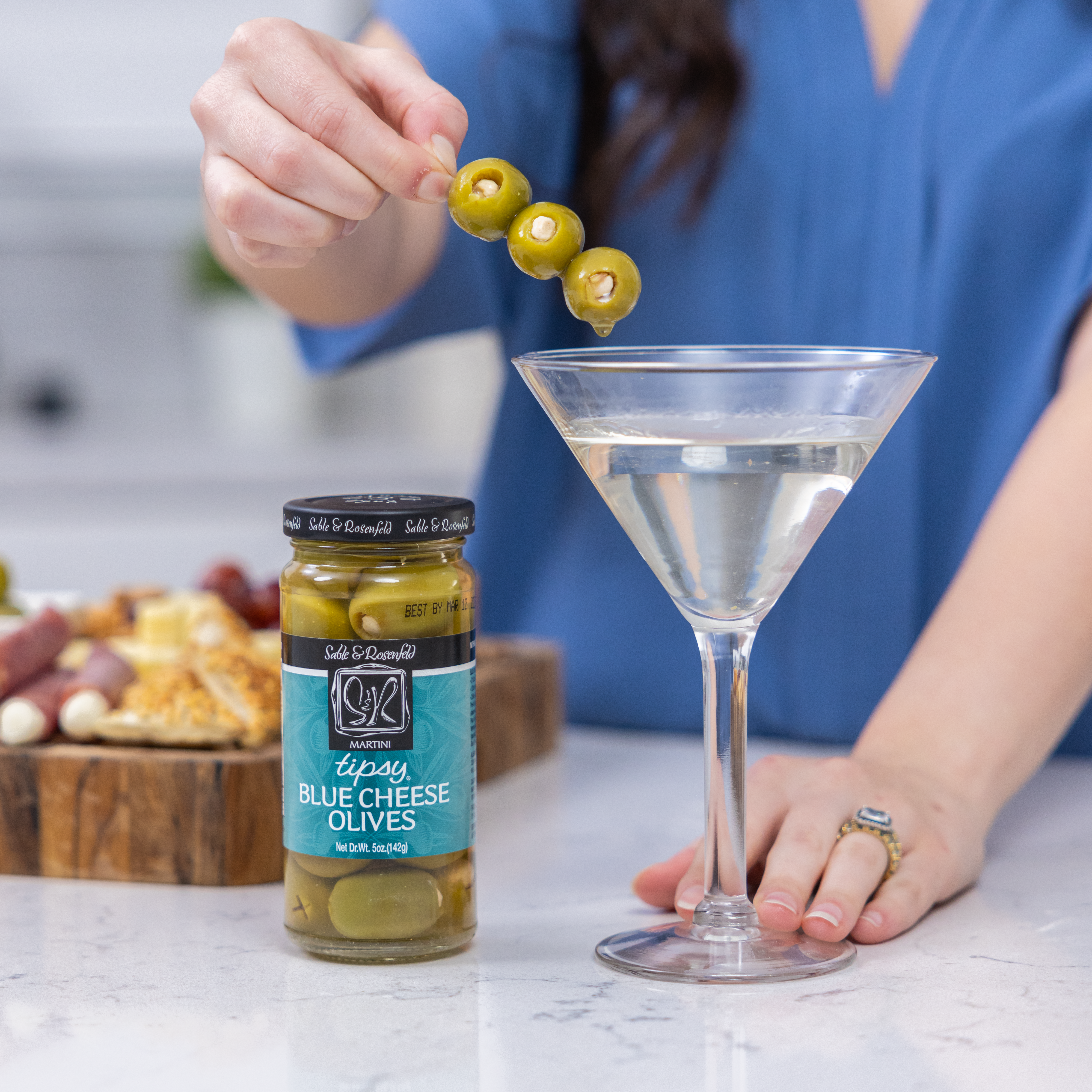 A hand garnishing a martini with skewered blue cheese olives, alongside a jar of Sable & Rosenfeld Blue Cheese Tipsy Olives and a charcuterie board in the background.