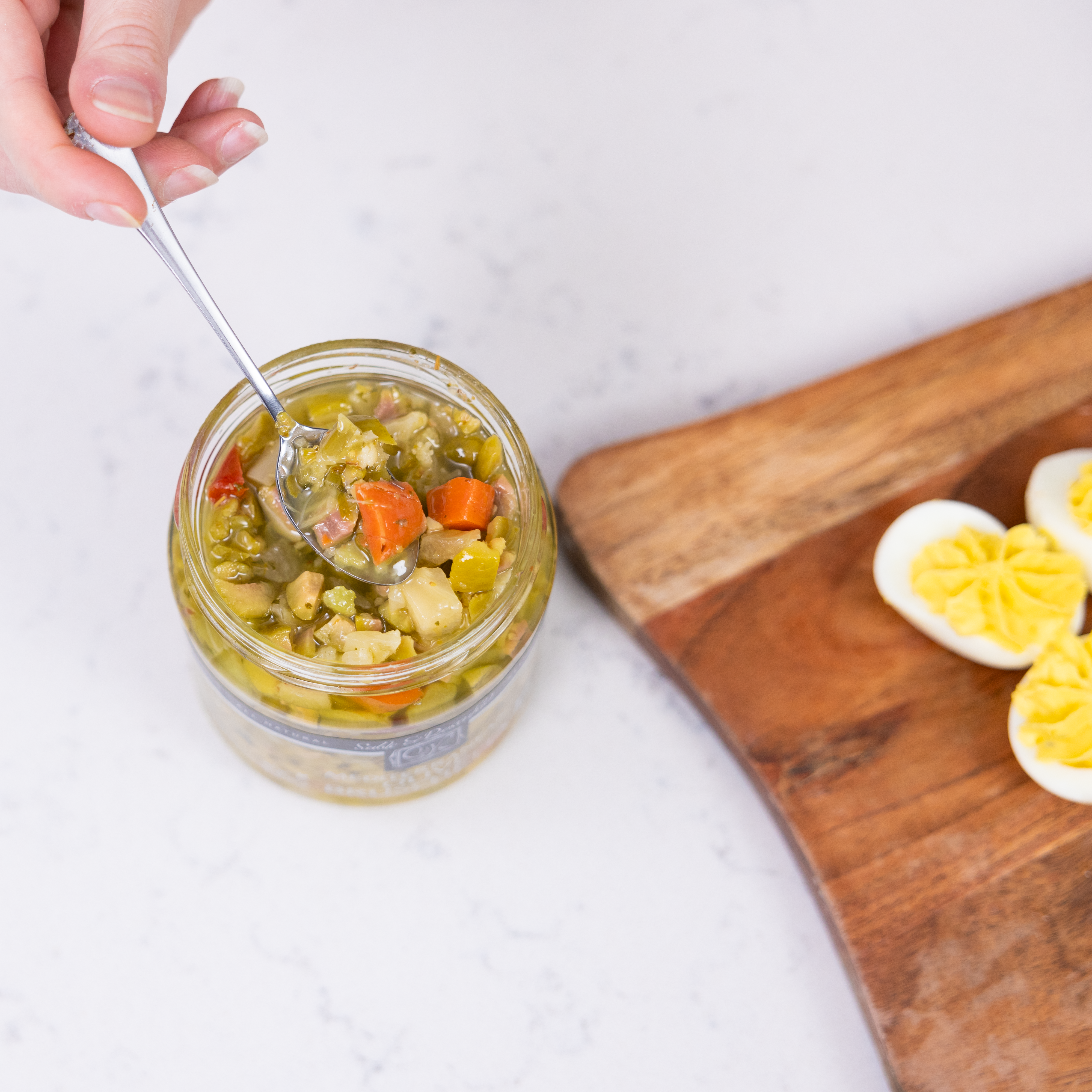 A hand scooping a spoonful of bruschetta olive mix from an open jar, with a wooden board featuring halved boiled eggs on a marble countertop.