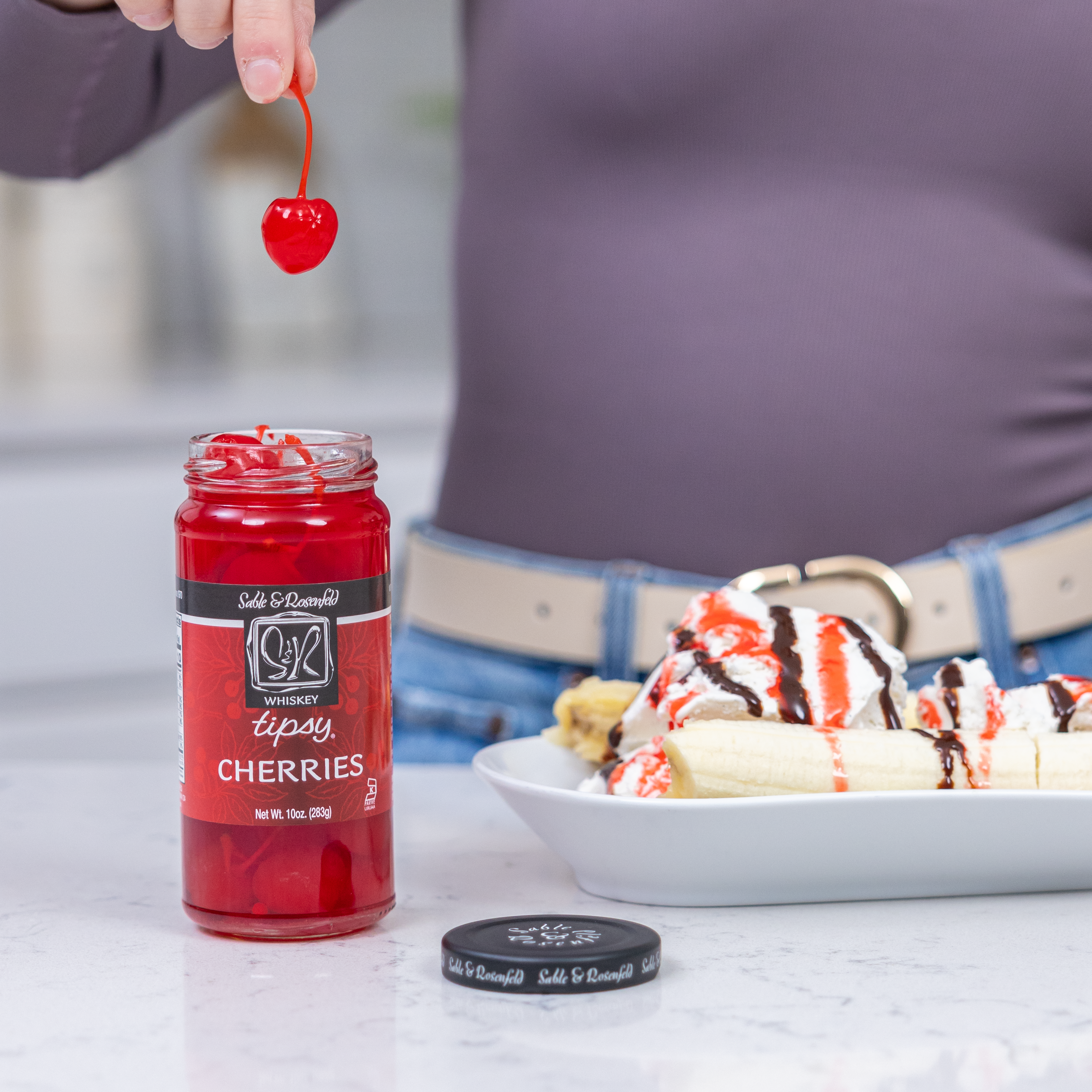 A jar of Sable & Rosenfeld Whiskey Tipsy Cherries on a white countertop, with a hand lifting a whiskey-soaked cherry. In the background, a banana split drizzled with chocolate and cherry syrup enhances the gourmet dessert experience.