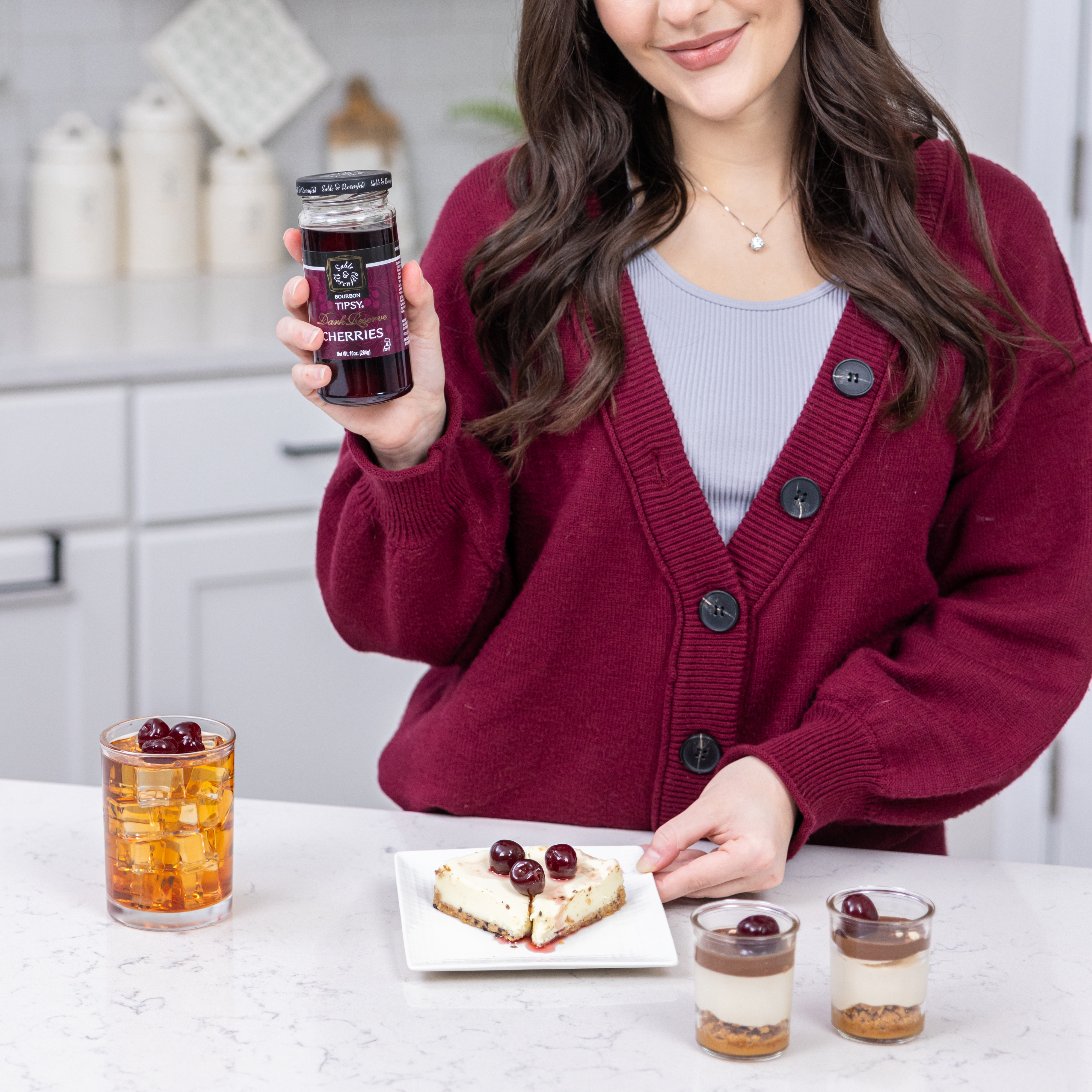 Woman holding a jar of cherries with cherry infused bourbon cocktail and desserts on a kitchen counter.