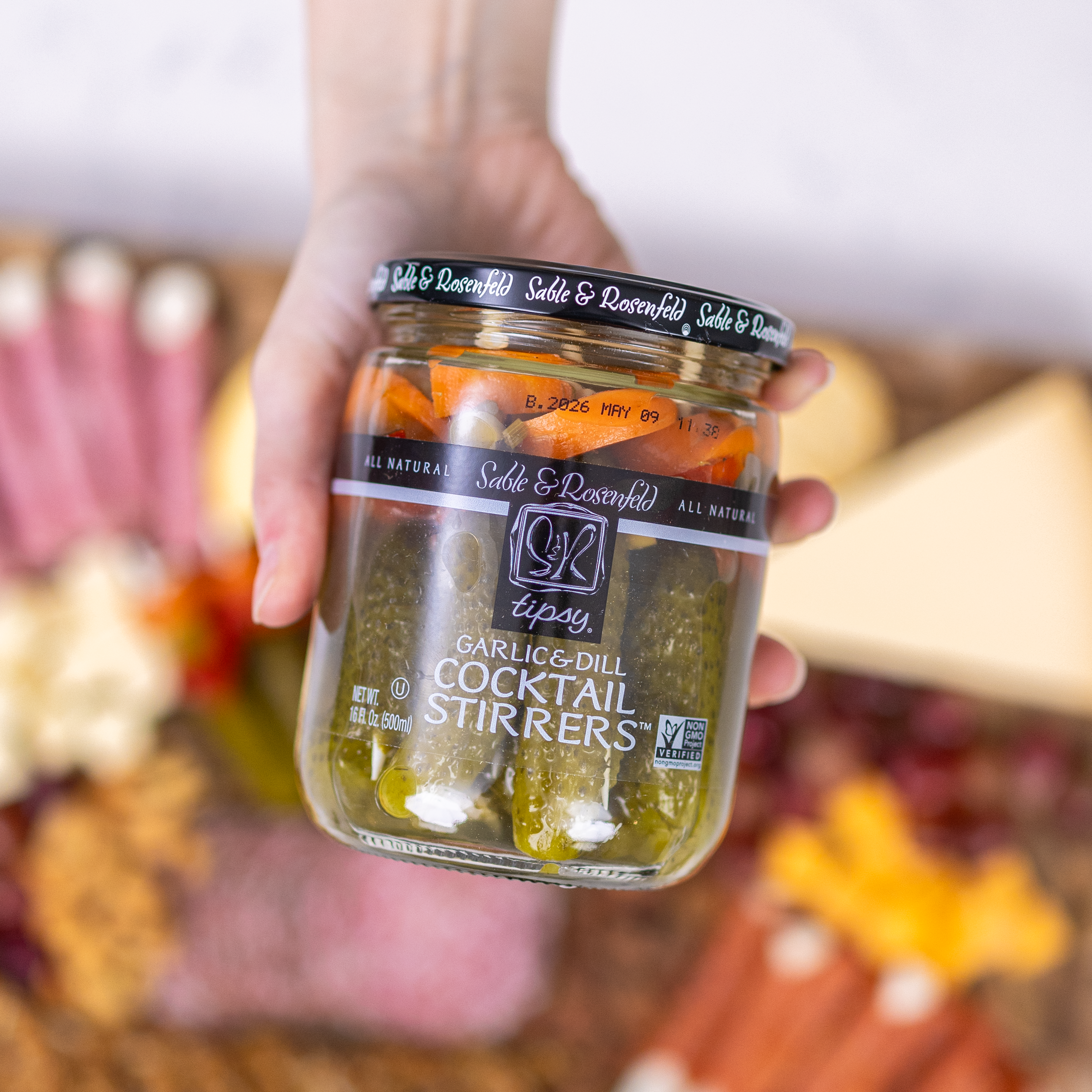 A hand holding a jar of Sable & Rosenfeld Tipsy Garlic & Dill Cocktail Stirrers, filled with pickled gherkins and vegetables, with a gourmet charcuterie board in the background.