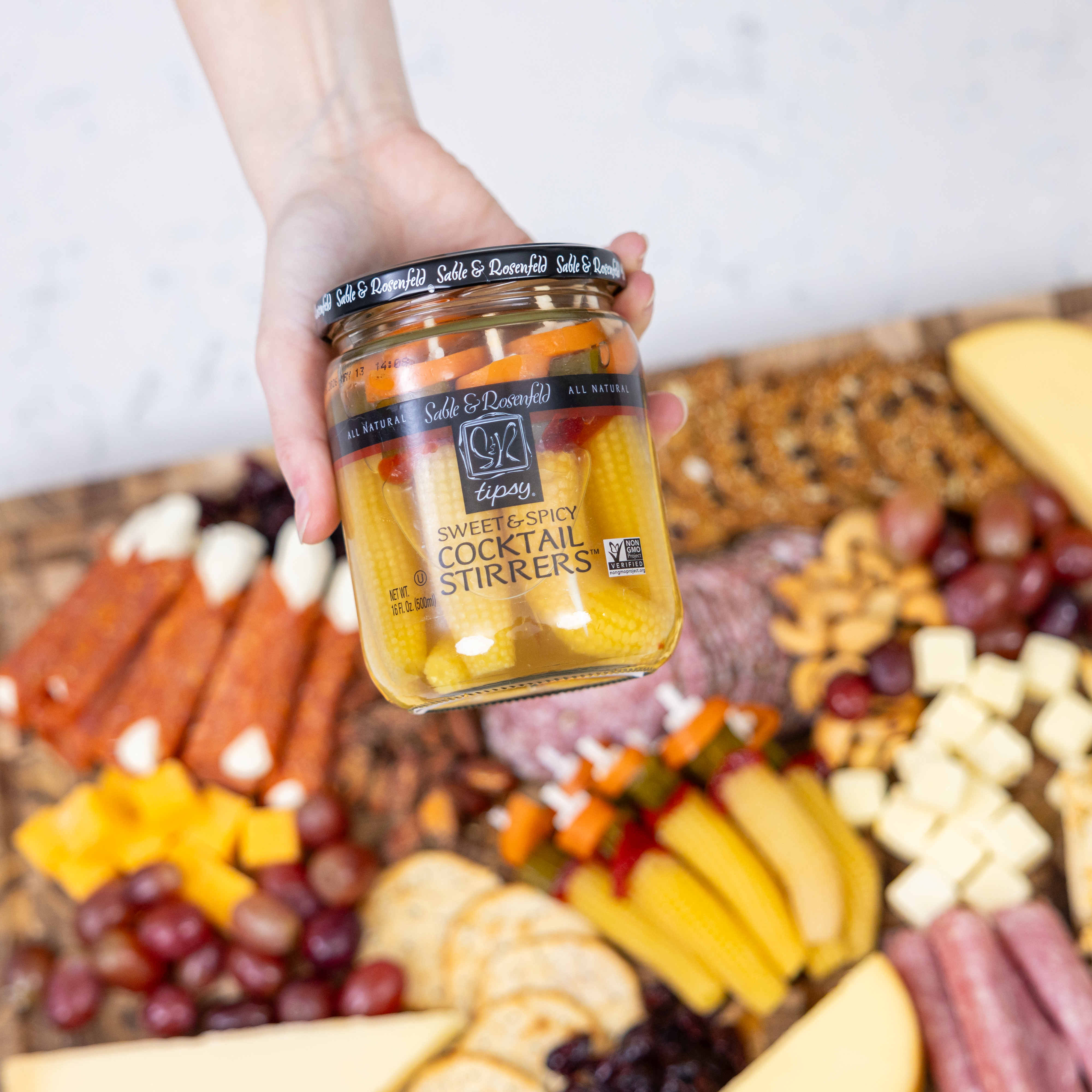 A hand holding a jar of Sable & Rosenfeld Sweet & Spicy Cocktail Stirrers, featuring corn skewers, pickles, and peppers, above a charcuterie board with cheese, meats, and crackers.