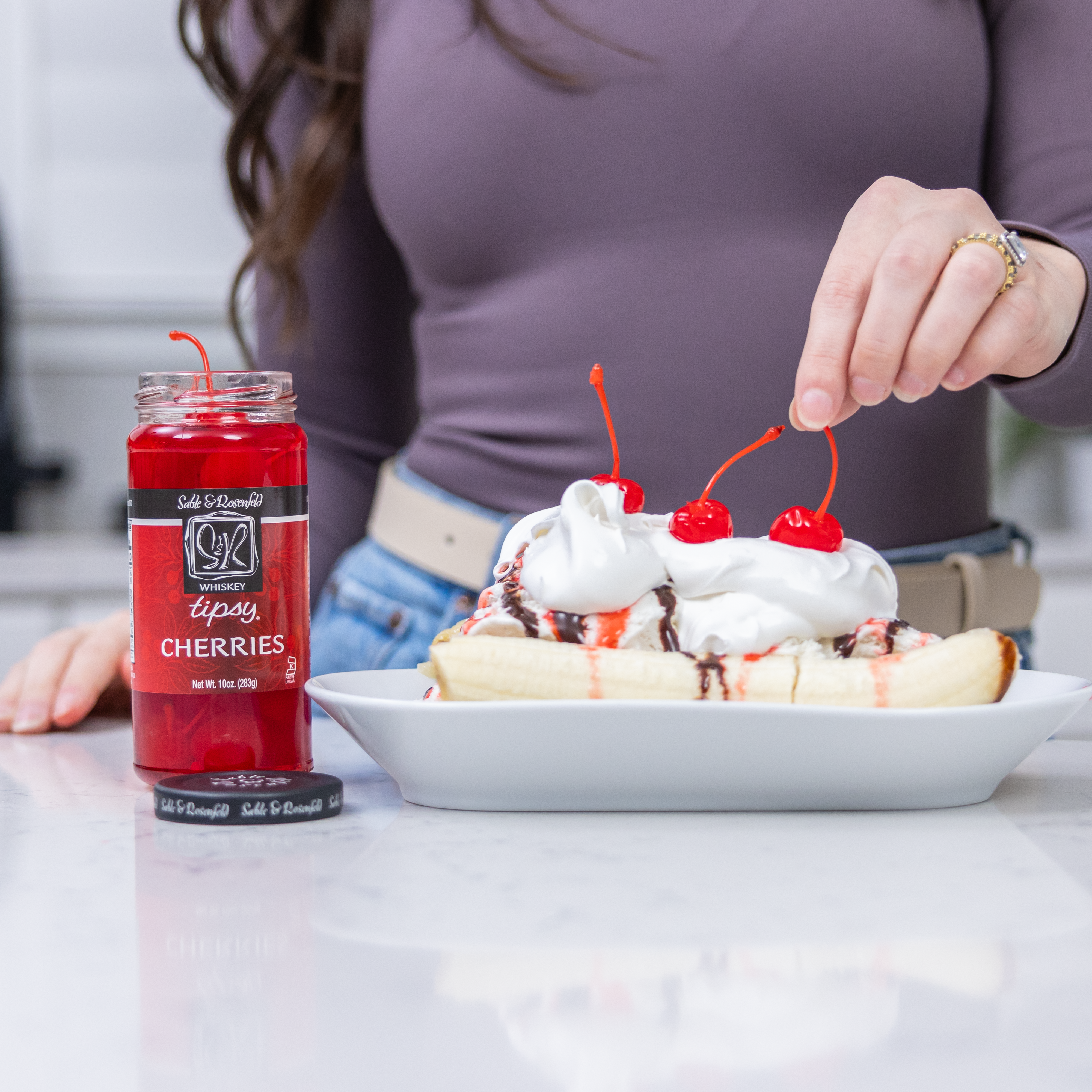 A gourmet banana split dessert topped with whipped cream, chocolate drizzle, and vibrant red whiskey-infused tipsy cherries. A person is delicately placing a cherry on top, with a jar of Tipsy Cherries by Sable & Rosenfeld prominently displayed on a sleek kitchen countertop.