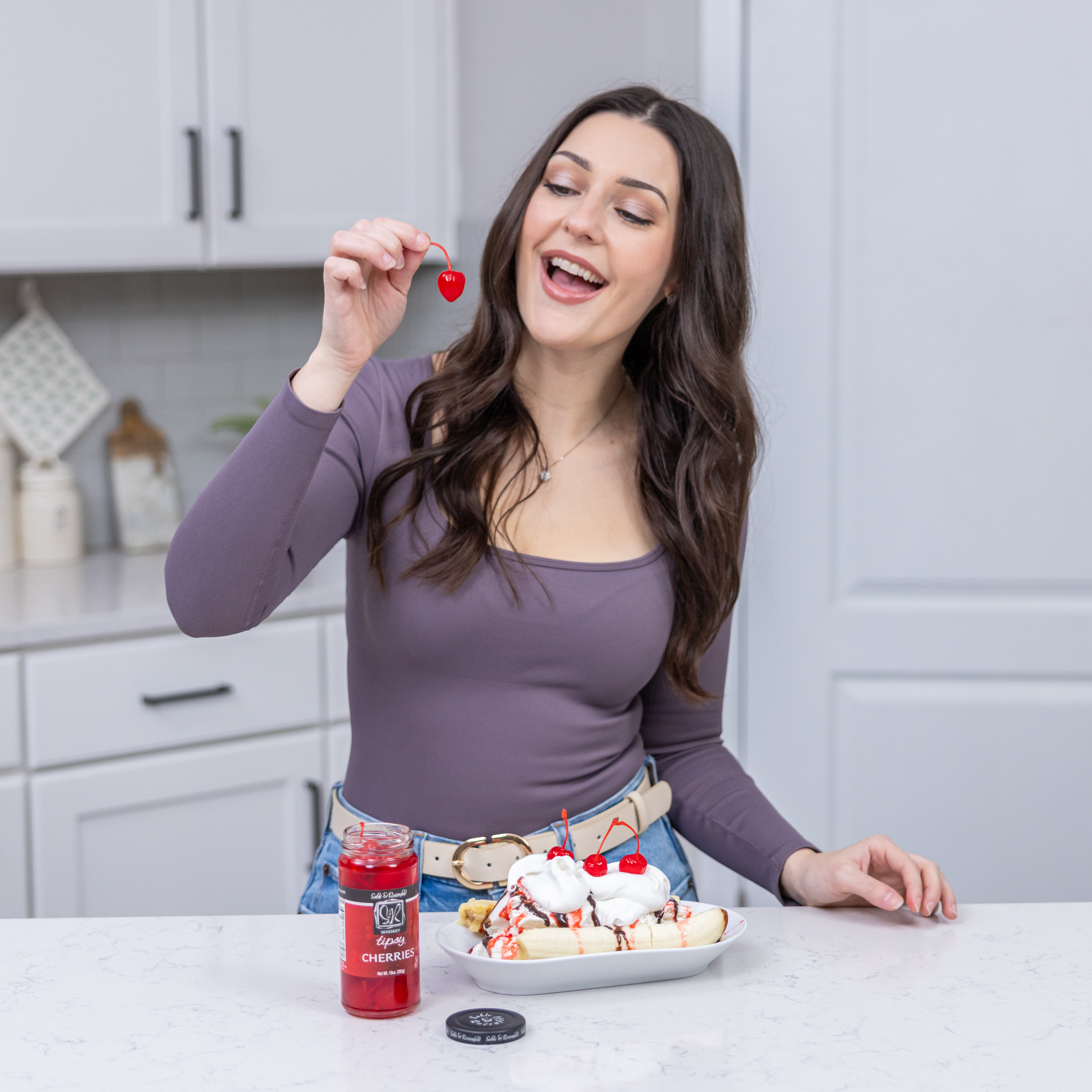 A smiling woman in a modern kitchen holding a whiskey-soaked cherry above a banana split topped with whipped cream, chocolate drizzle, and cherries. A jar of Sable & Rosenfeld Whiskey Tipsy Cherries sits on the countertop, making for a perfect gourmet food gift.