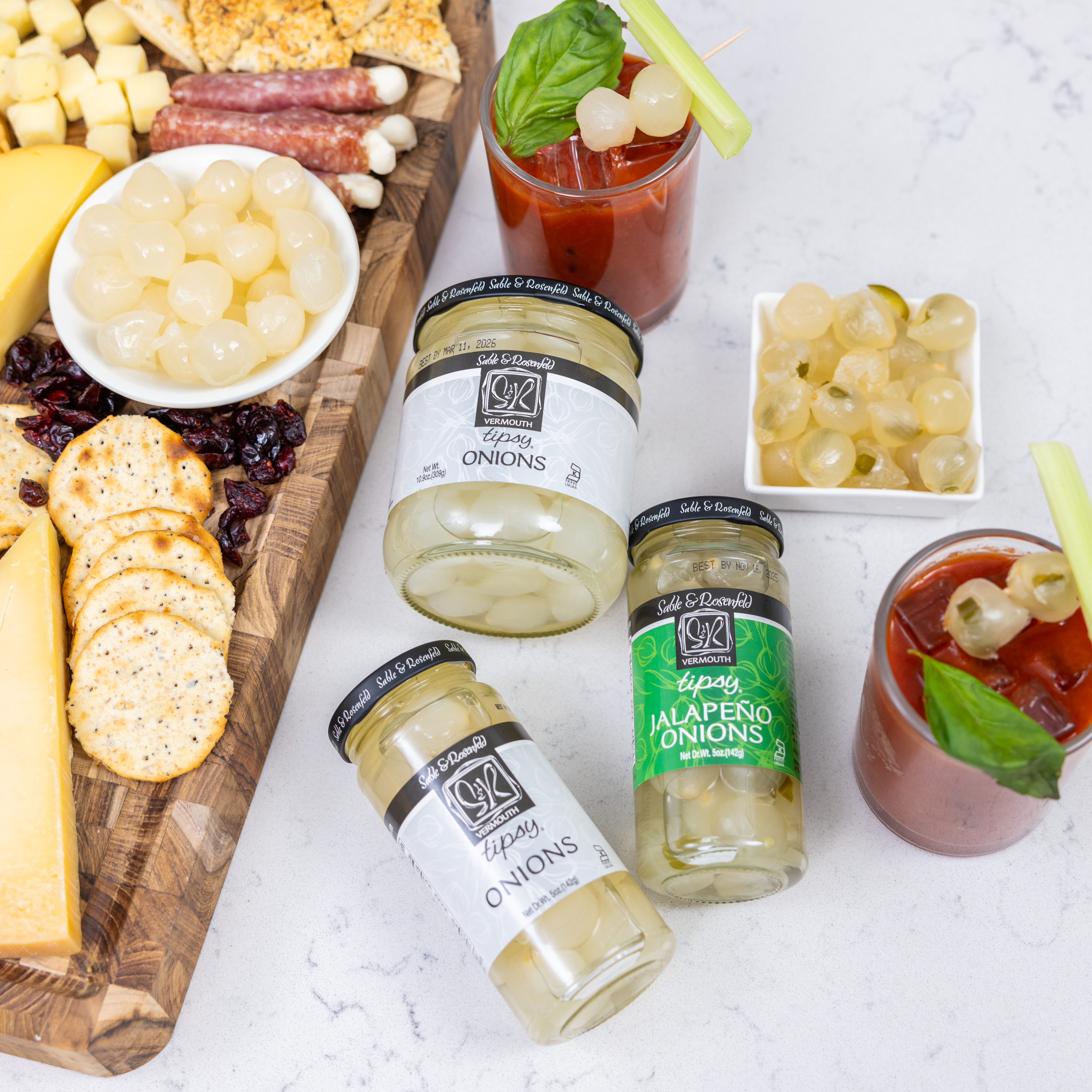 An assortment of Sable & Rosenfeld gourmet food store products, including Tipsy Onions and Jalapeño Onions, displayed alongside a charcuterie board with cheese, crackers, and Bloody Mary cocktails garnished with onions and celery.