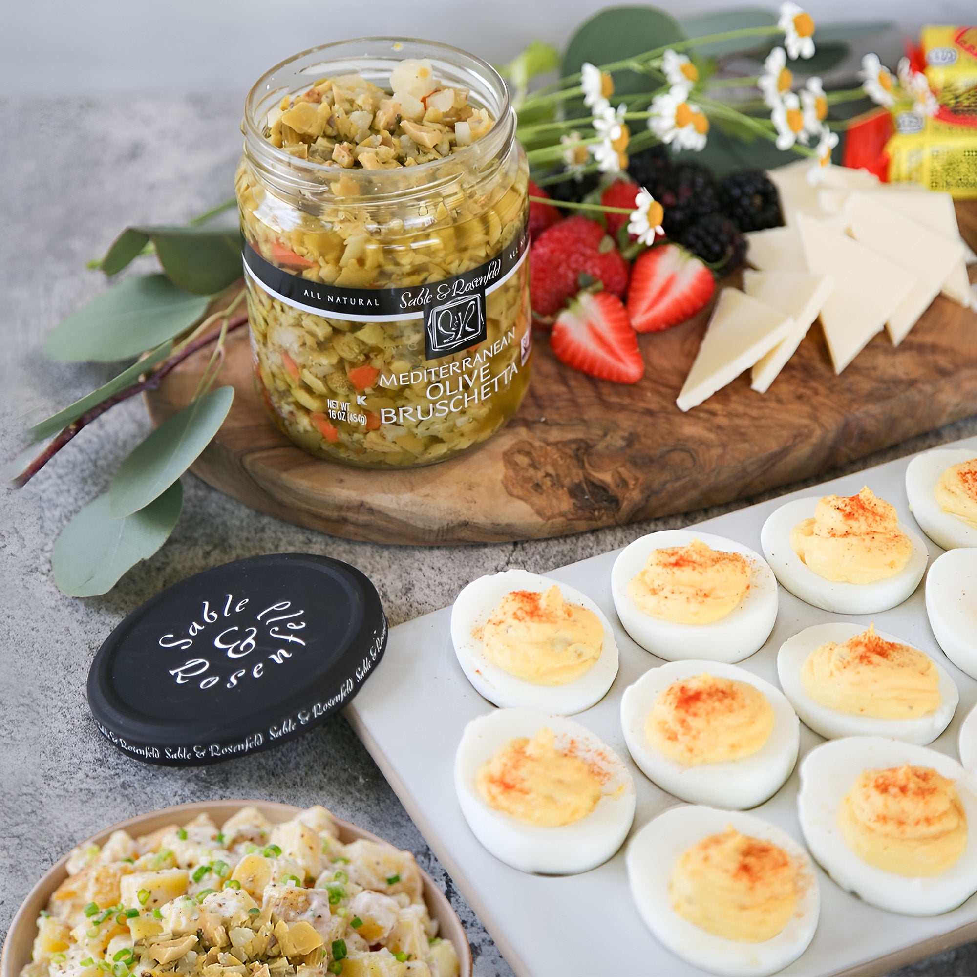 A jar of Sable & Rosenfeld Mediterranean Green Olive Bruschetta on a wooden serving board, surrounded by deviled eggs, fresh berries, cheese slices, and a pasta salad.