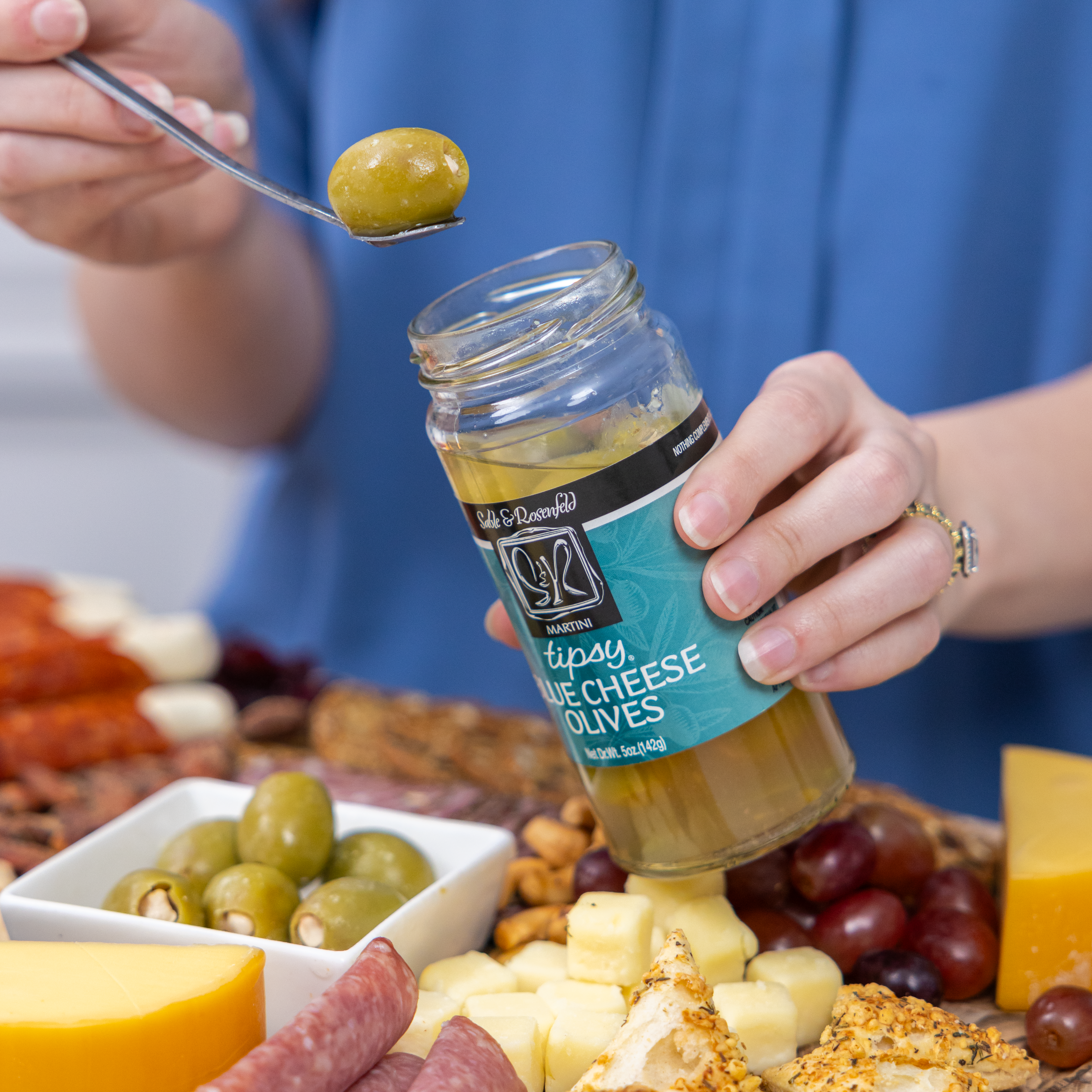 A person holding a jar of Sable & Rosenfeld Tipsy Blue Cheese Olives while picking up a green olive stuffed with blue cheese, with a charcuterie board featuring cheese, grapes, and nuts in the background.