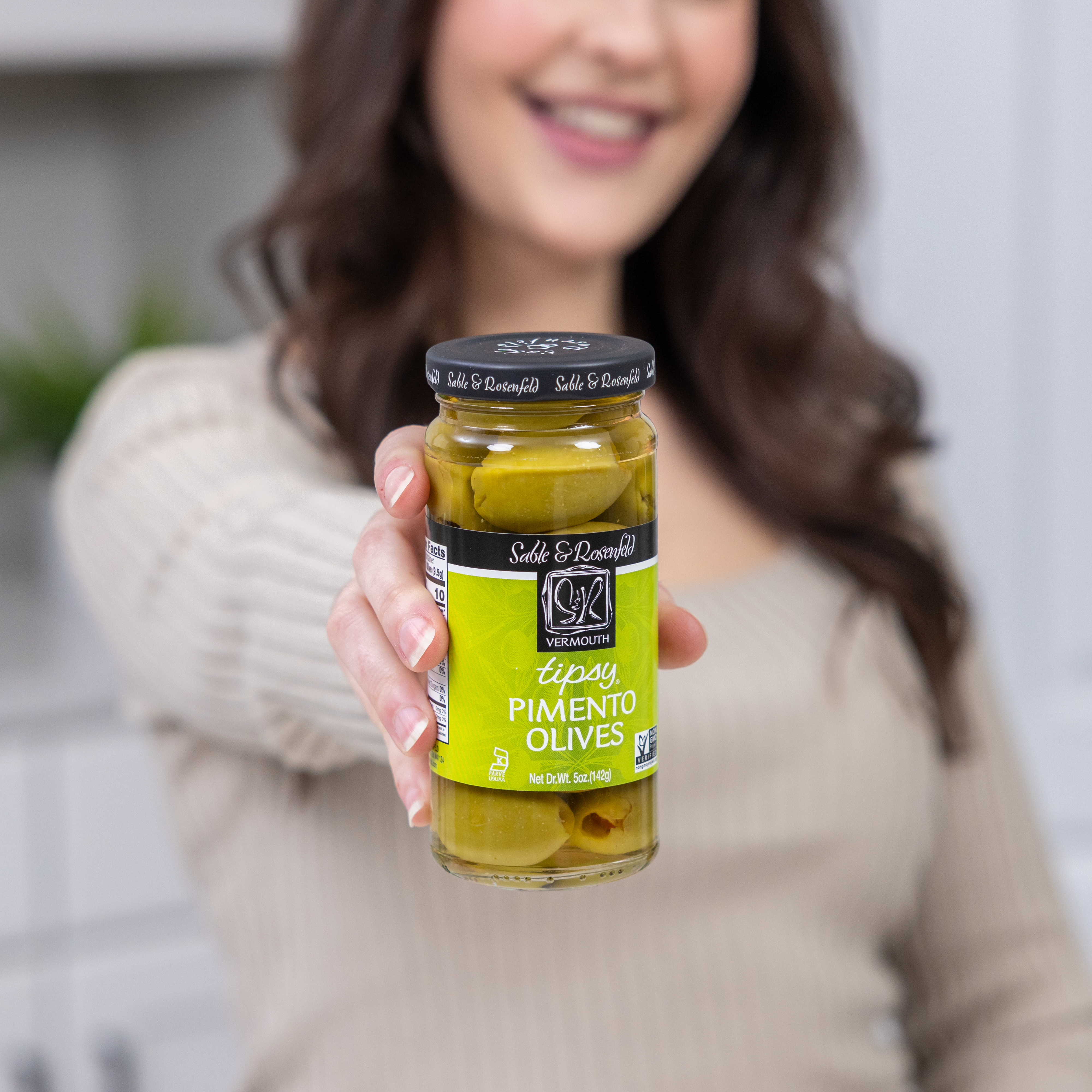 A smiling woman holding a jar of Sable & Rosenfeld Tipsy Pimento Olives, showcasing green olives stuffed with pimentos in a clear glass jar with a black lid and green label.