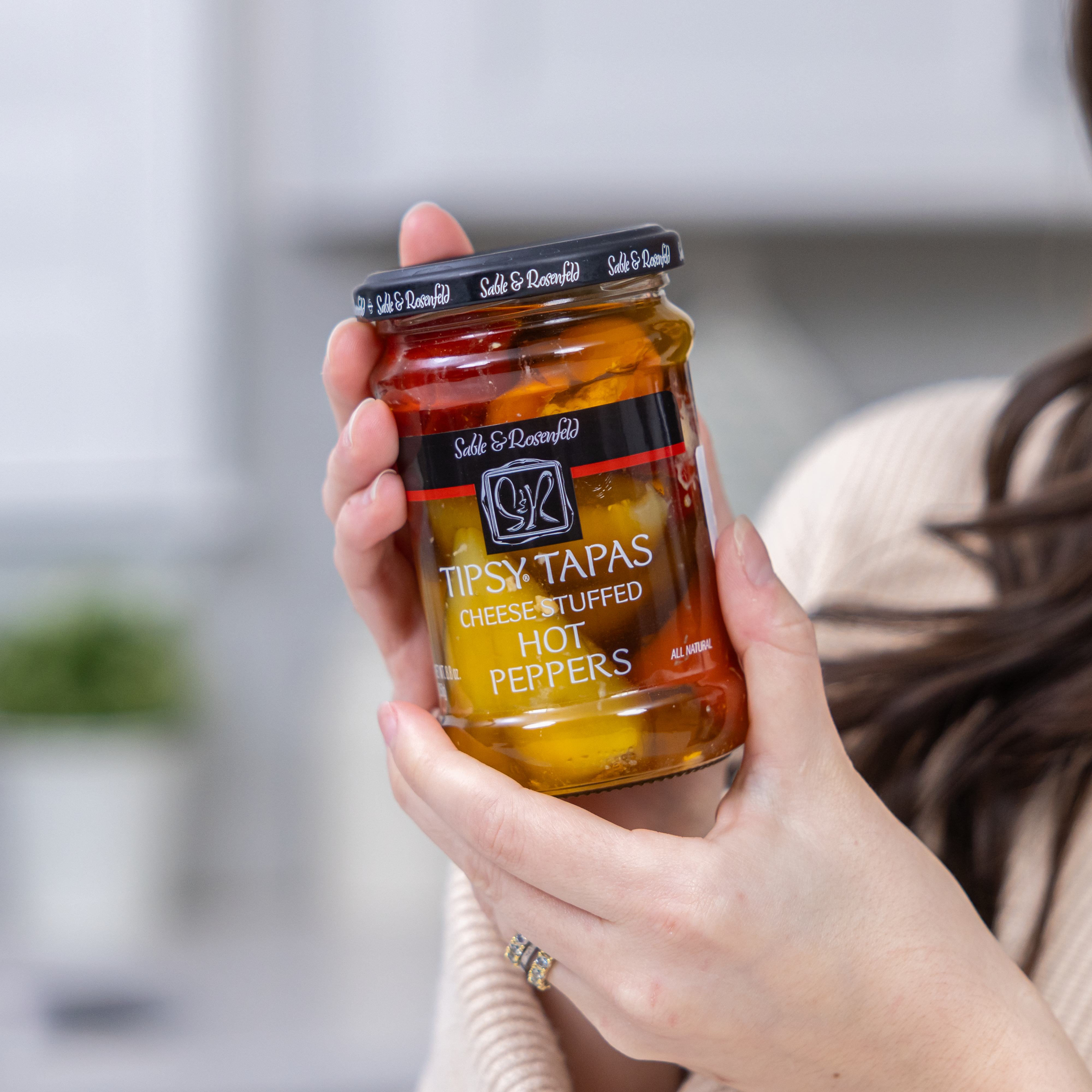 A woman holding a jar of Sable & Rosenfeld Tipsy Tapas Hot Peppers stuffed with cream cheese, featuring vibrant red and yellow peppers in brine.