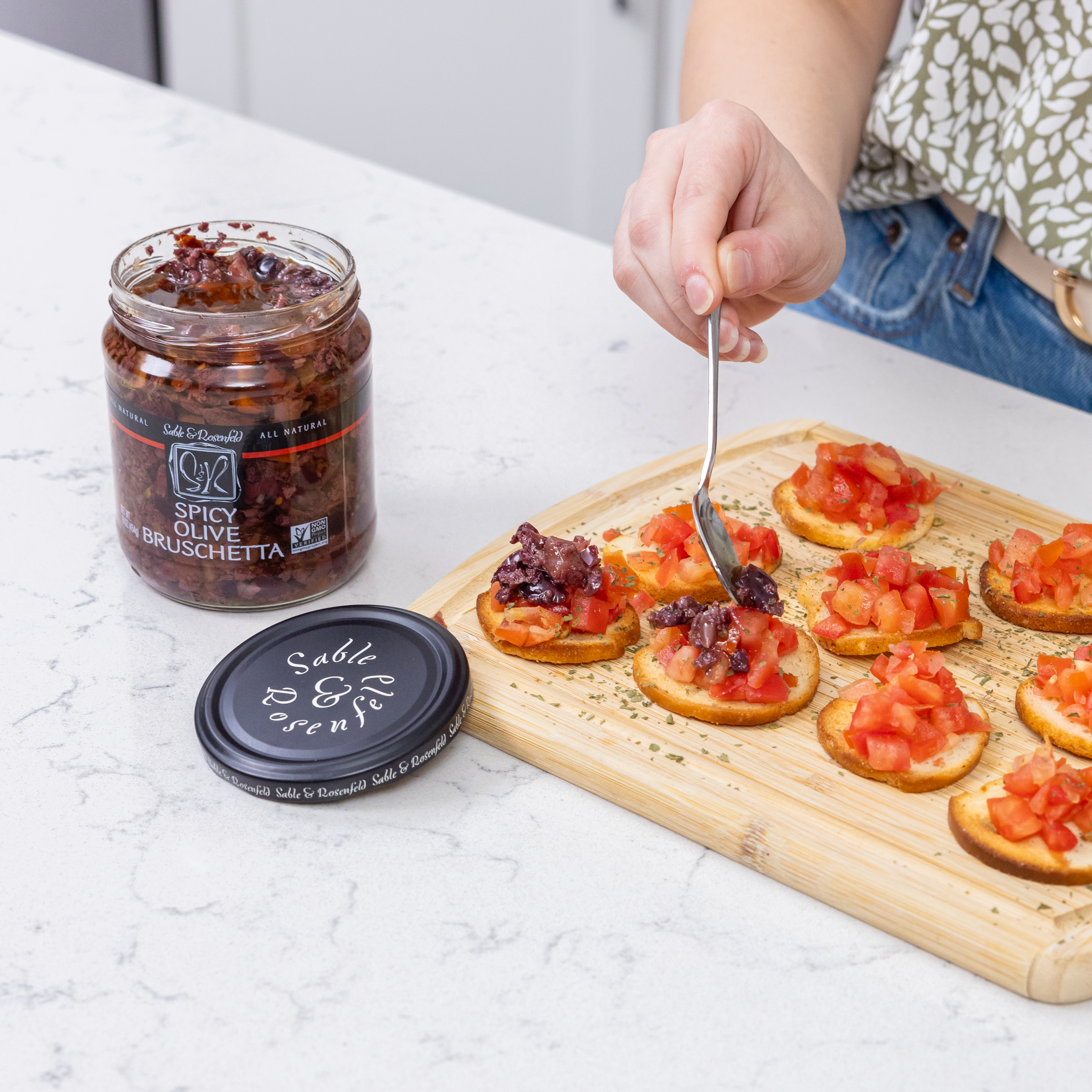 A person spooning Spicy Olive Bruschetta onto crispy toasted bread rounds with diced tomatoes, served on a wooden board. A jar of Spicy Olive Bruschetta is placed beside the bruschetta on a white kitchen counter.